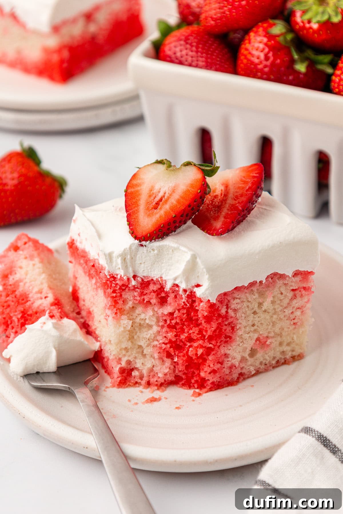 A slice of strawberry poke cake with white frosting and two halved strawberries on top sits on a plate with a fork. In the background, there is a container filled with fresh strawberries, inviting you to indulge.