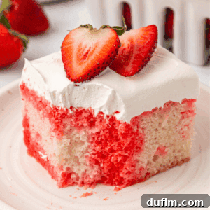 A slice of strawberry poke cake with white frosting, topped with two fresh strawberry halves, sits on a white plate. Fresh strawberries are visible in the background, a perfect summer dessert.