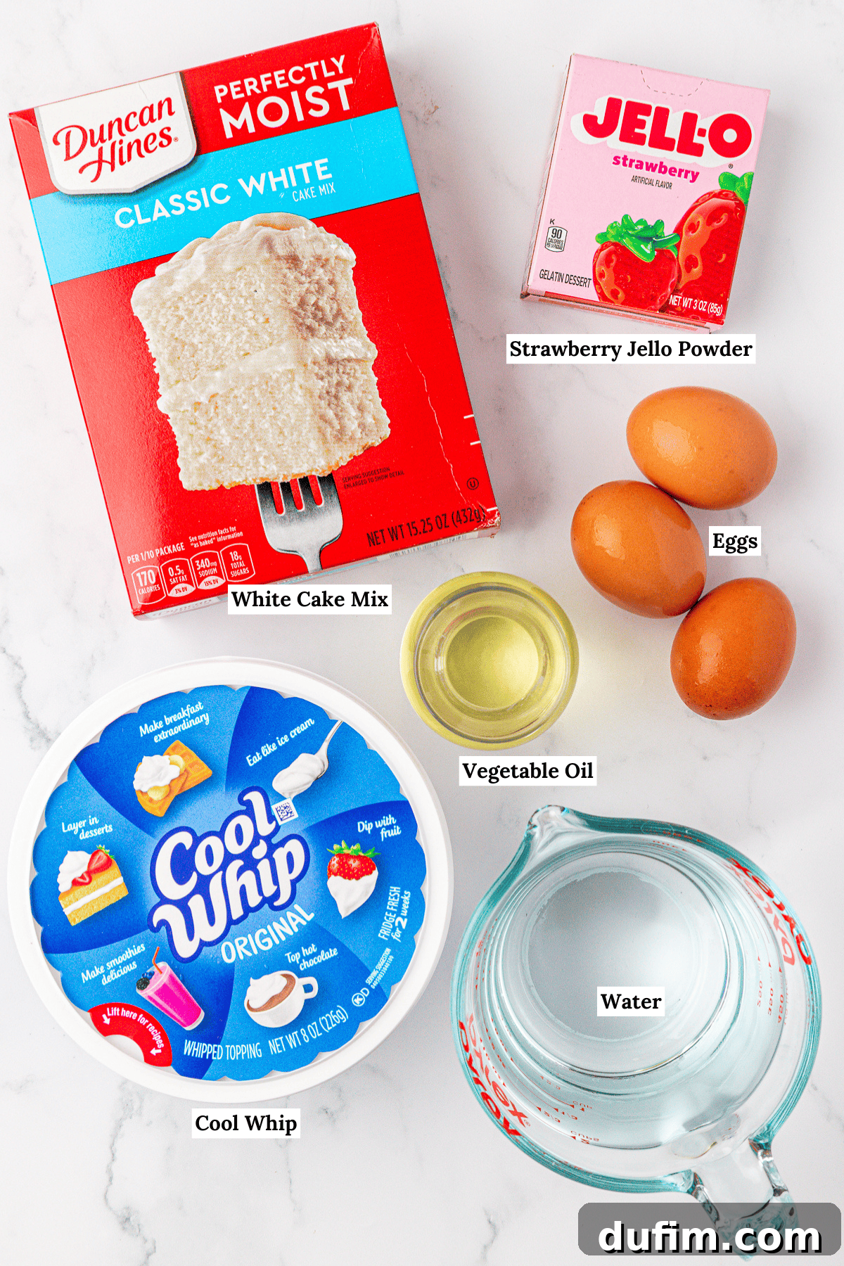Top-down view of ingredients for a dessert: white cake mix, strawberry Jello powder, three eggs, vegetable oil in a small bowl, Cool Whip container, and a measuring cup of water on a white marble surface, ready for baking.