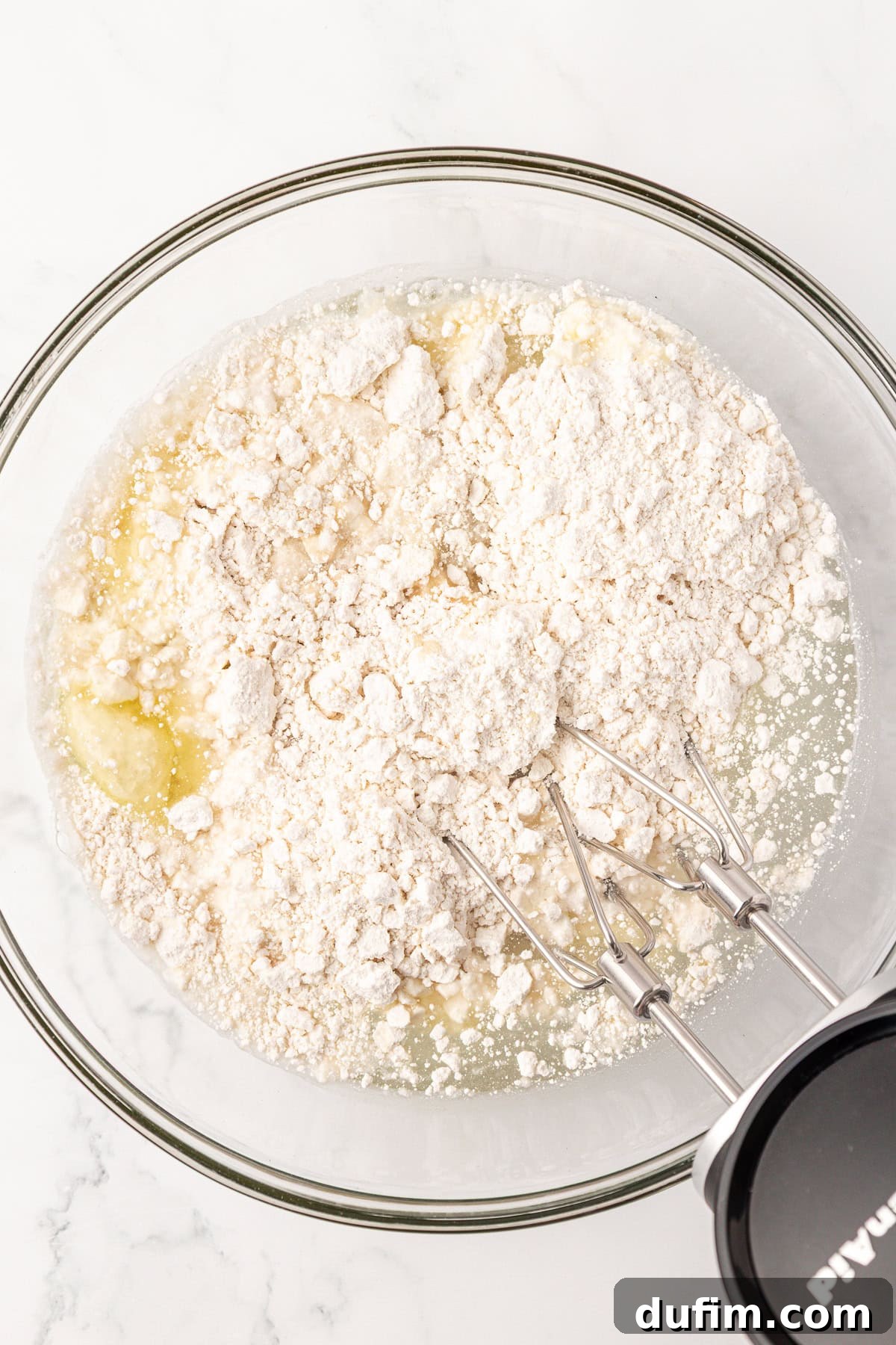 A glass bowl containing flour and wet ingredients is being mixed with an electric hand mixer on a white surface, creating a smooth cake batter.