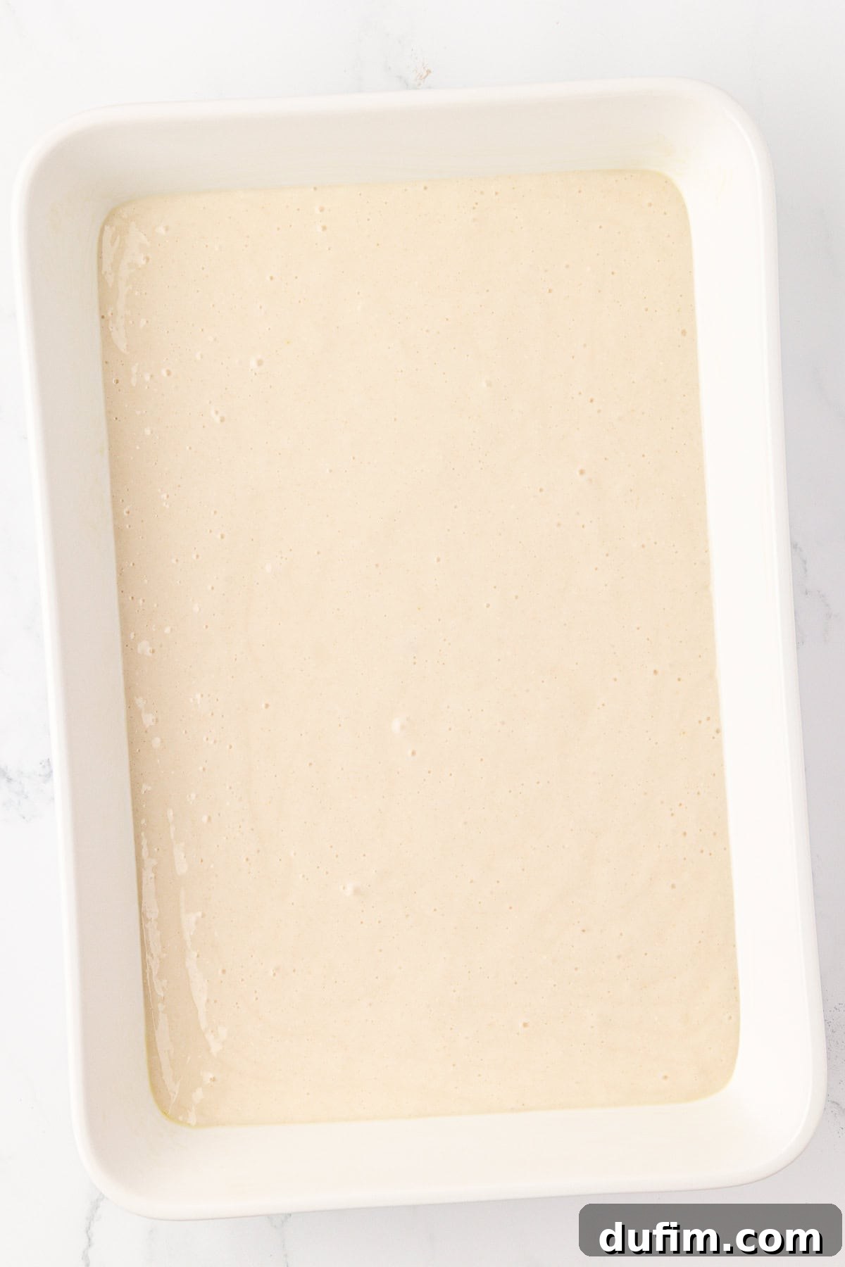 A white rectangular baking dish filled with an even layer of light beige cake batter, set on a white marble surface, ready for baking.