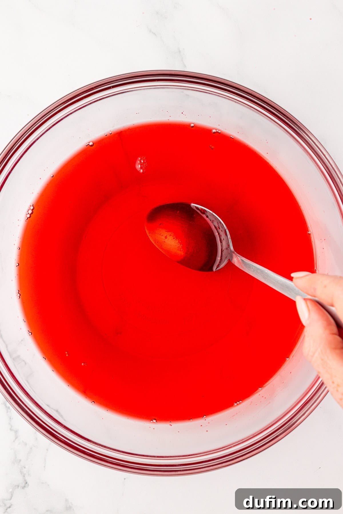 A hand holding a spoon stirs bright red liquid in a clear glass bowl on a white surface, preparing the vibrant Jello mixture.