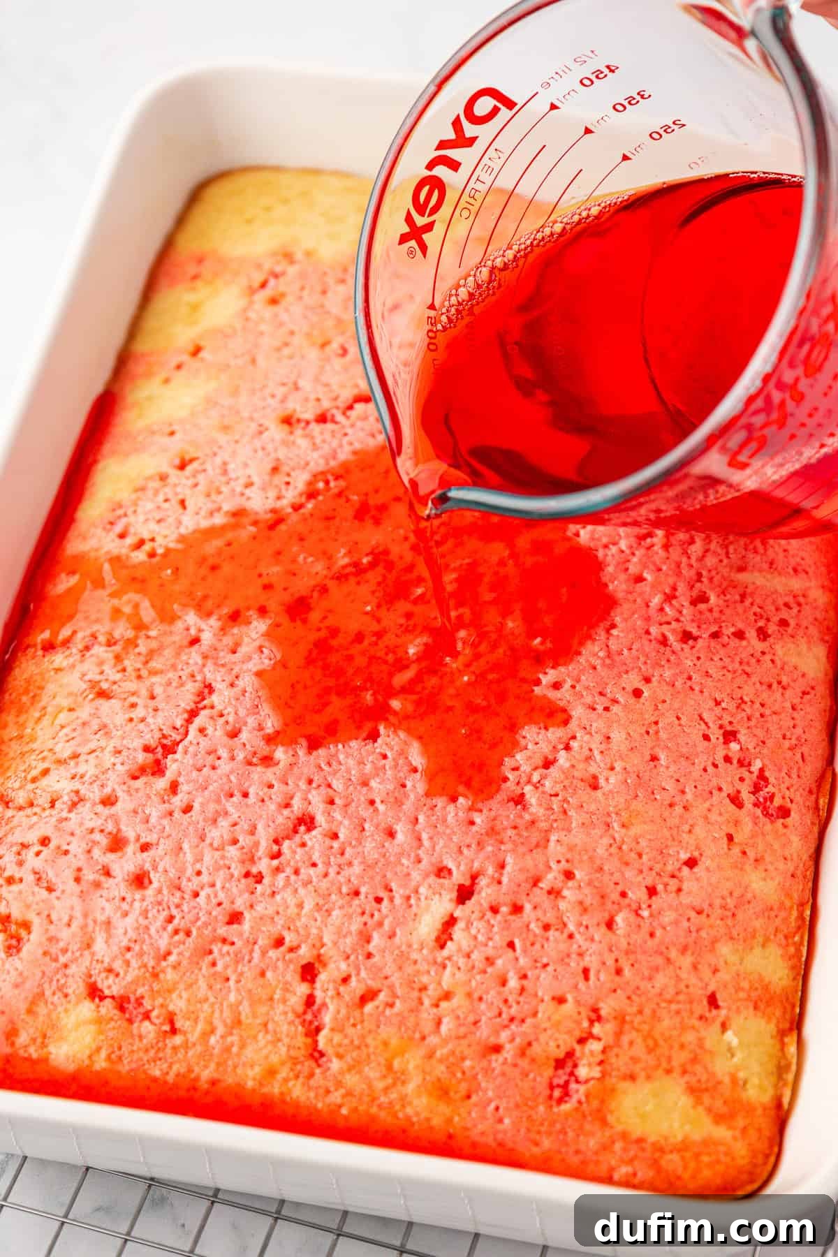 A glass measuring cup pours red liquid over a rectangular yellow cake in a white baking dish, with the cake showing small holes where the liquid is being absorbed, creating a beautiful marbling effect.