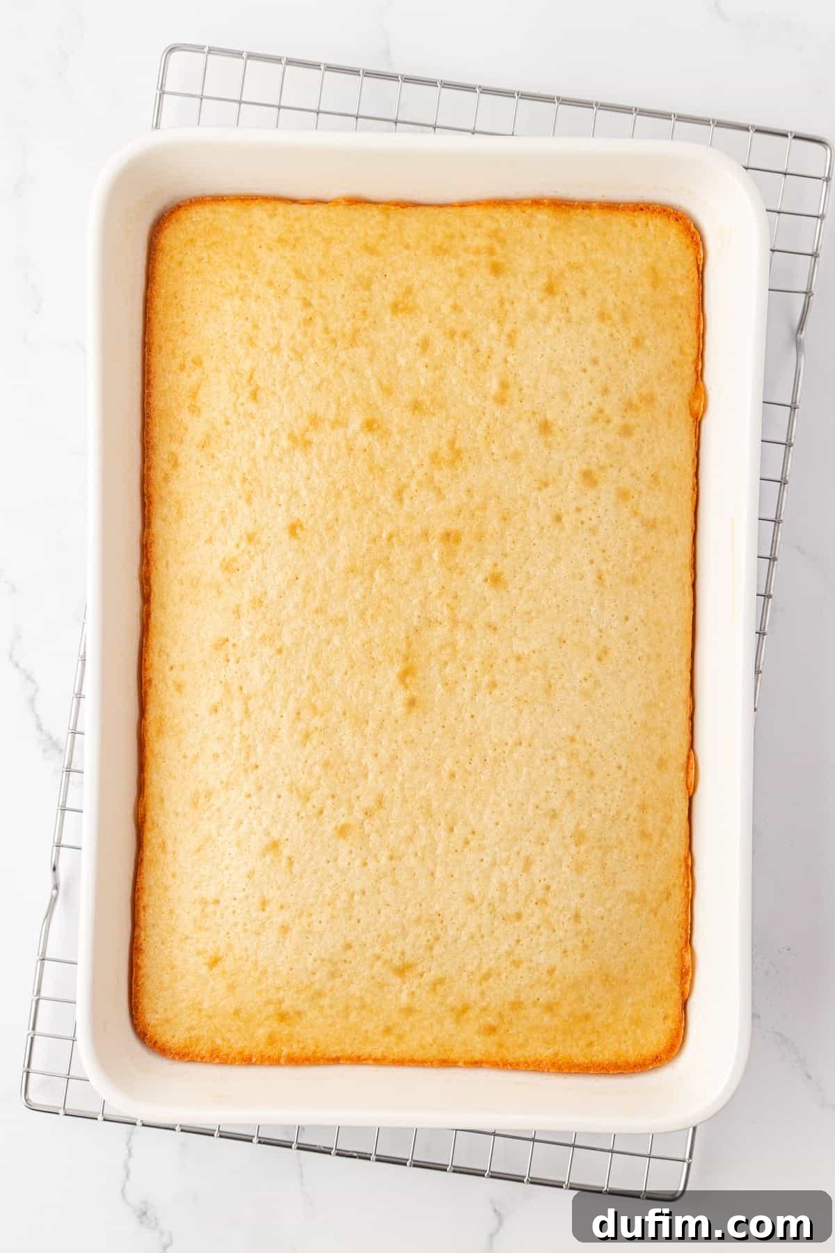 A rectangular vanilla sheet cake in a white baking dish, resting on a metal cooling rack atop a white marble surface. The cake has a golden-brown edge and a smooth, lightly textured top, cooling to perfection.