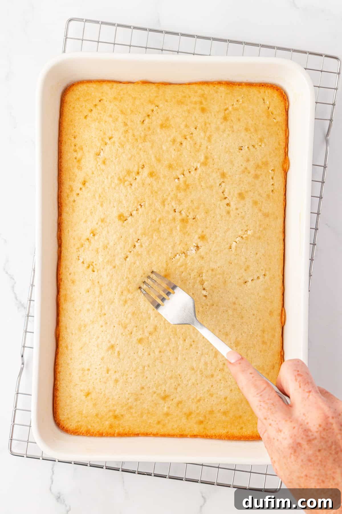 A hand uses a fork to poke holes in a rectangular, golden-brown cake still in a white baking dish, which rests on a metal cooling rack over a marble surface, preparing it for the Jello infusion.
