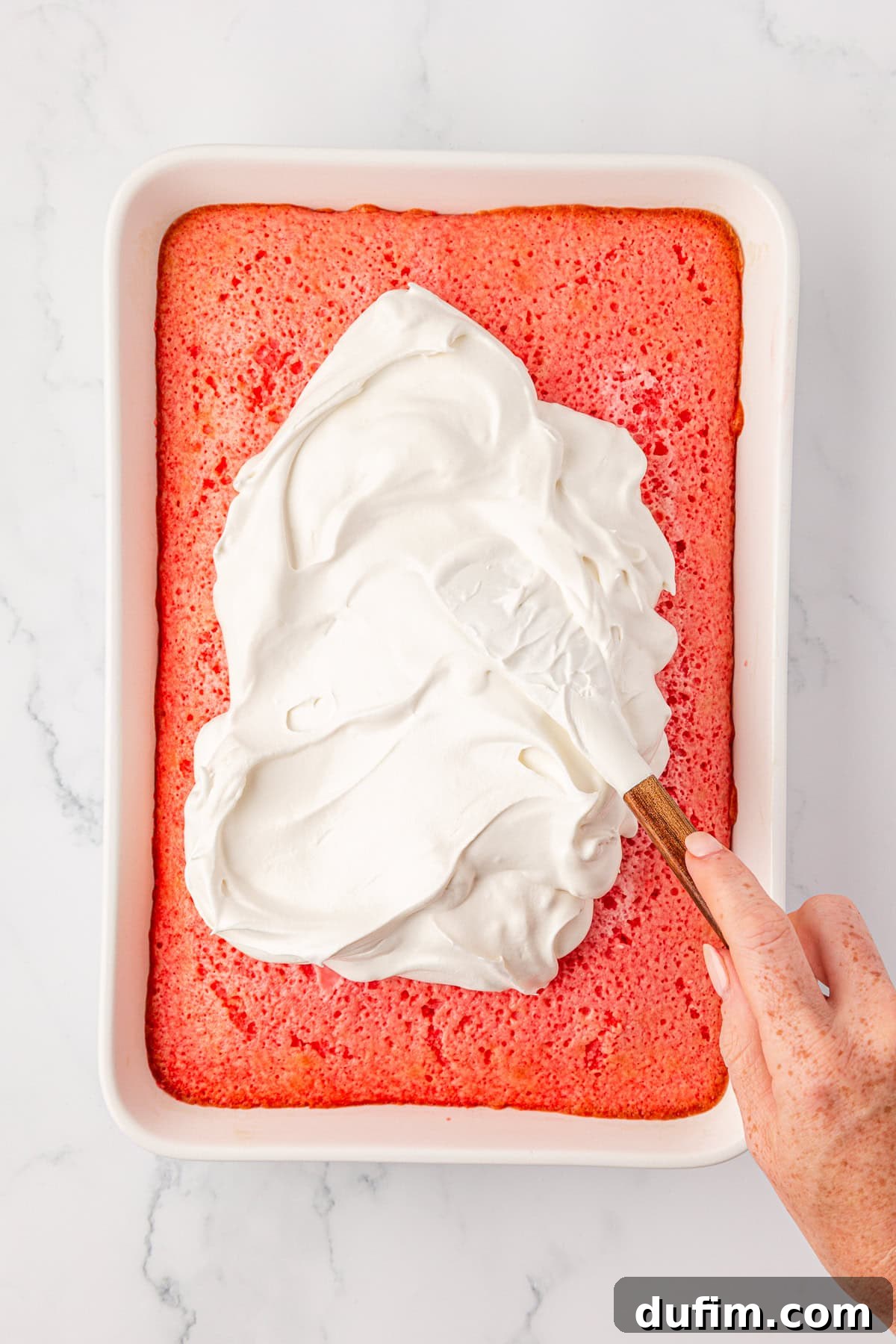 A hand spreads a thick layer of white whipped topping evenly over a rectangular pink cake in a white baking dish on a marble surface, preparing the final layer.