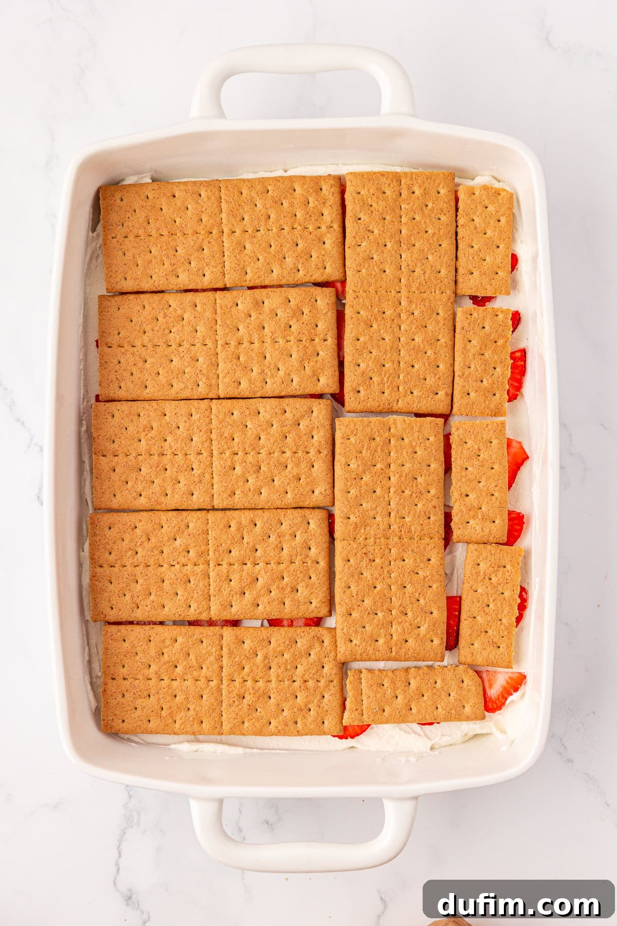 No-Bake Strawberry Dream 11 A white baking dish filled with a dessert layer of graham crackers on top of cream and sliced strawberries, sitting on a marble surface.