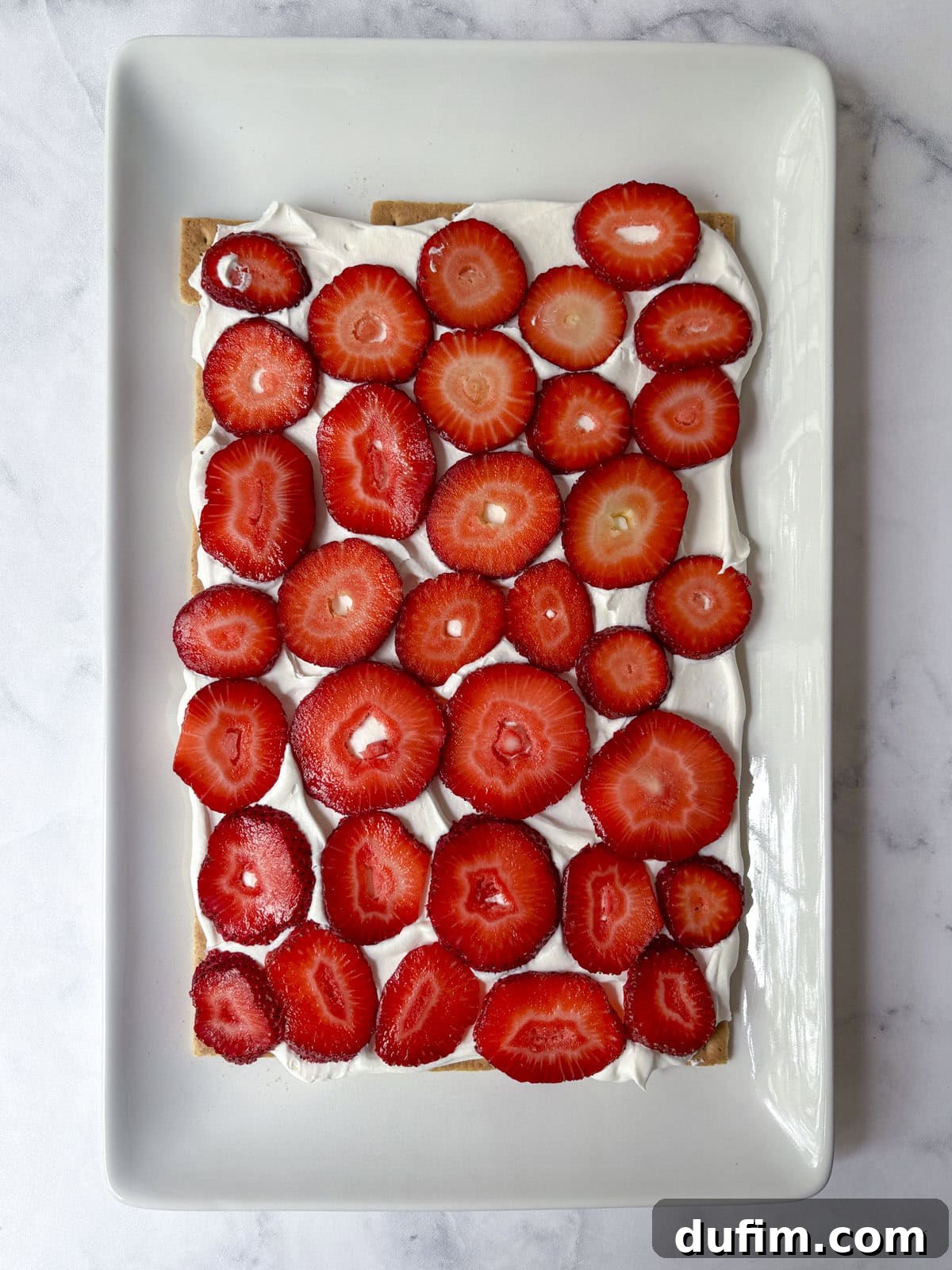 No-Bake Strawberry Dream 12 Rectangular dessert with a layer of whipped cream topped with neatly arranged fresh strawberry slices on a rectangular white plate, set on a light marble surface.