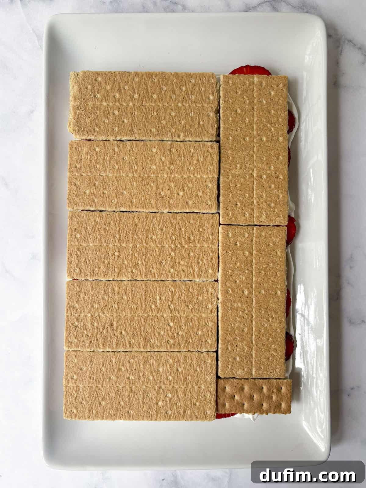 No-Bake Strawberry Dream 13 A rectangular white tray filled with neatly arranged graham cracker sheets covering a dessert with visible layers of strawberries and cream peeking out from the side.