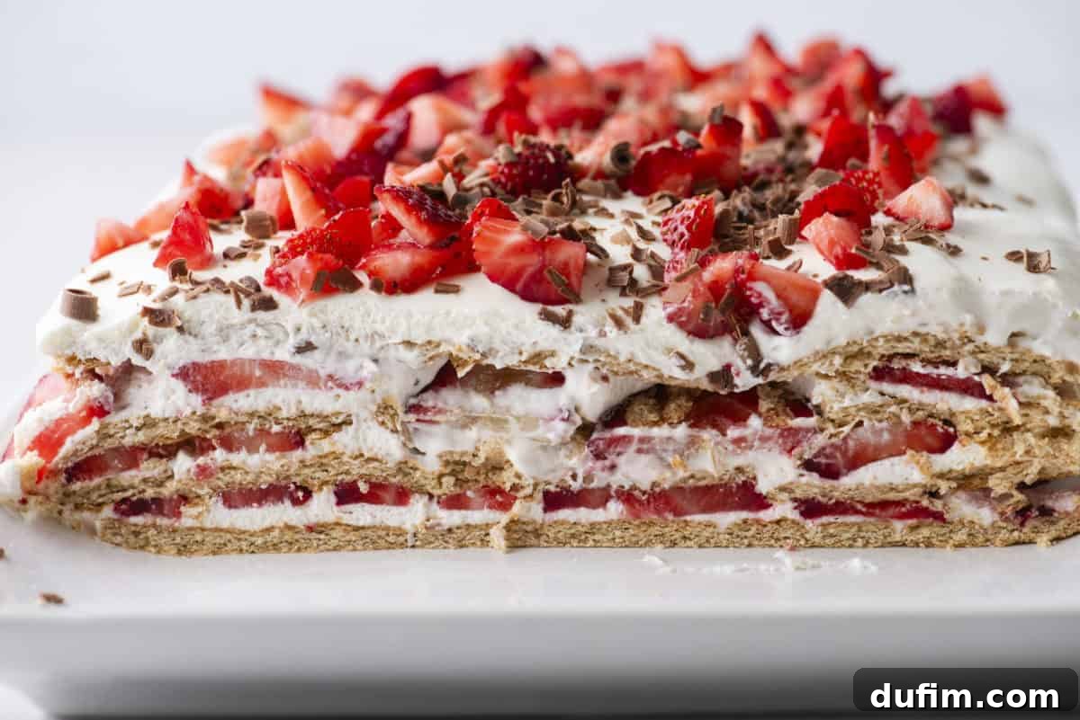 No-Bake Strawberry Dream 14 A rectangular layered dessert with whipped cream, sliced strawberries, graham crackers, and chocolate shavings, shown with a slice removed to reveal the layers inside.