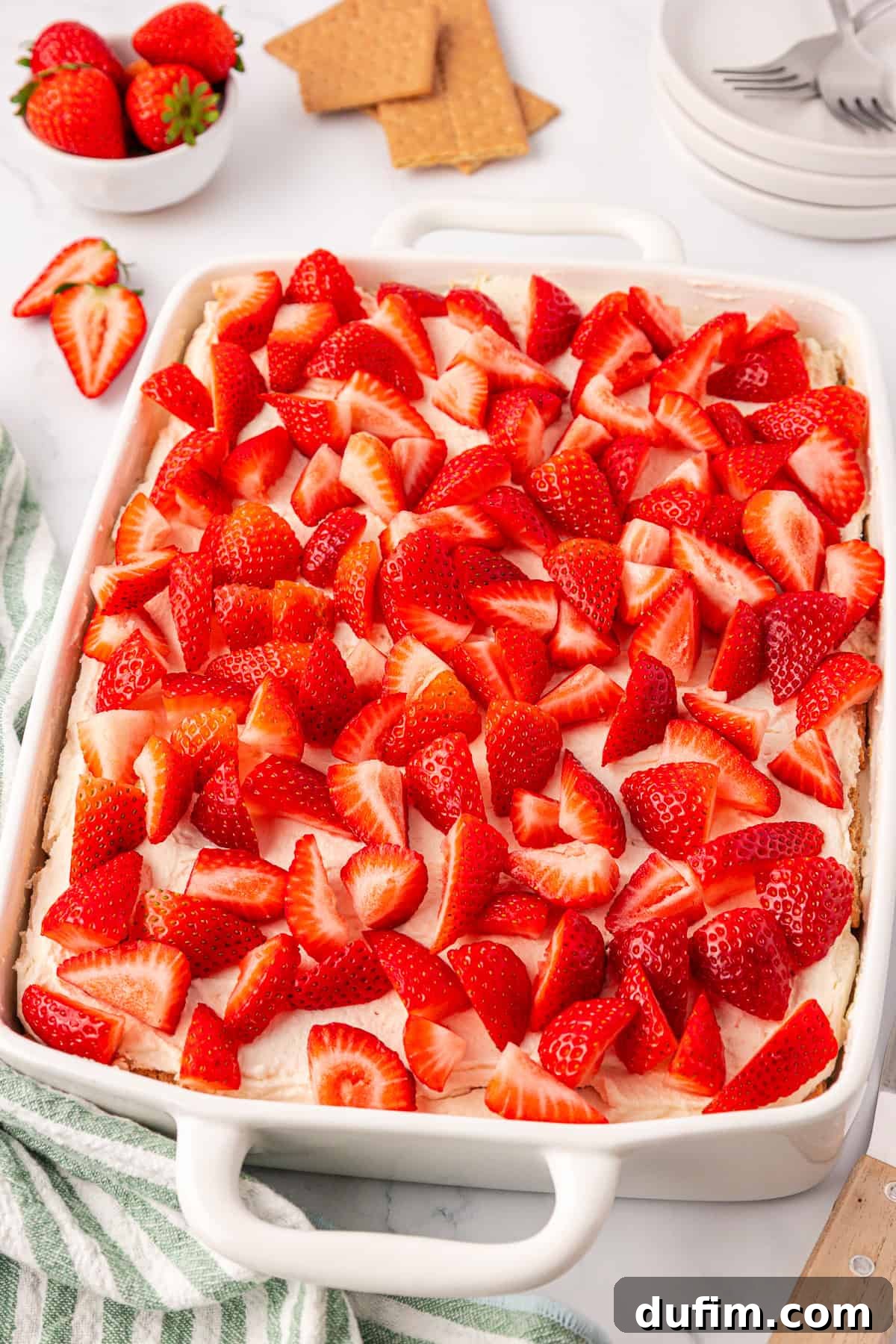 No-Bake Strawberry Dream 15 A white baking dish filled with a creamy dessert topped generously with sliced fresh strawberries. Nearby are plates with forks, whole strawberries, graham crackers, and a green-striped kitchen towel.