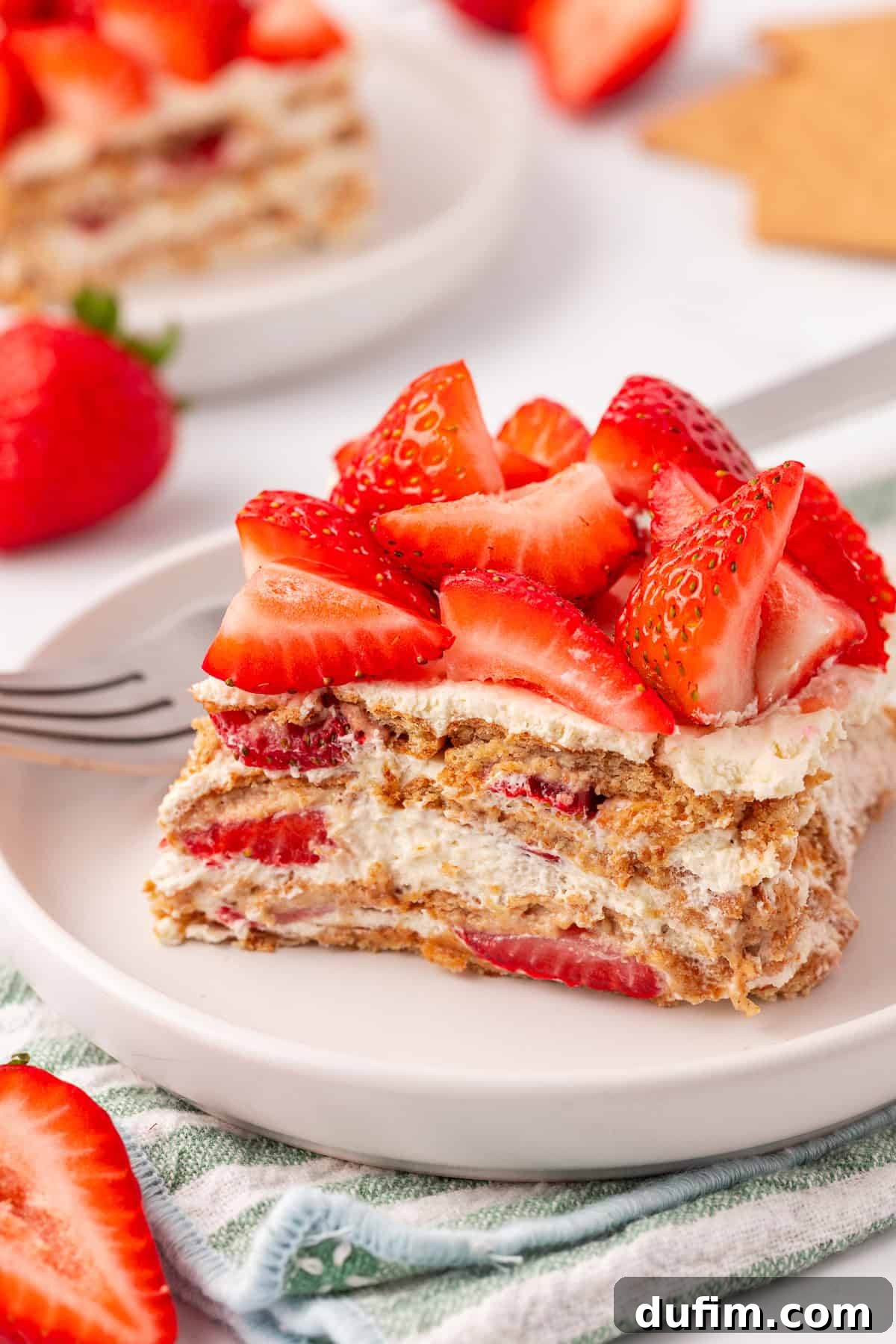 No-Bake Strawberry Dream 16 A slice of strawberry icebox cake with layers of whipped cream, graham crackers, and fresh strawberries on a white plate, garnished with strawberry pieces. A fork and additional strawberries are nearby.
