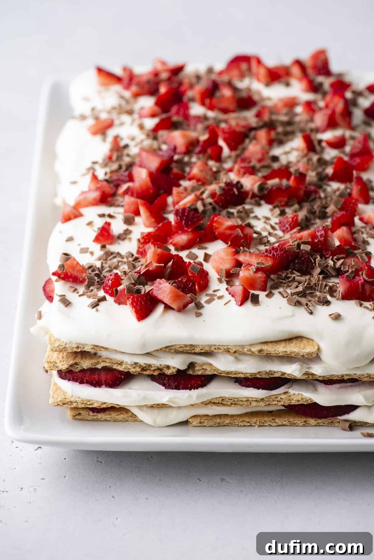 No-Bake Strawberry Dream 3 A layered dessert made with graham crackers, whipped cream, sliced strawberries, and chocolate shavings on a white rectangular plate.