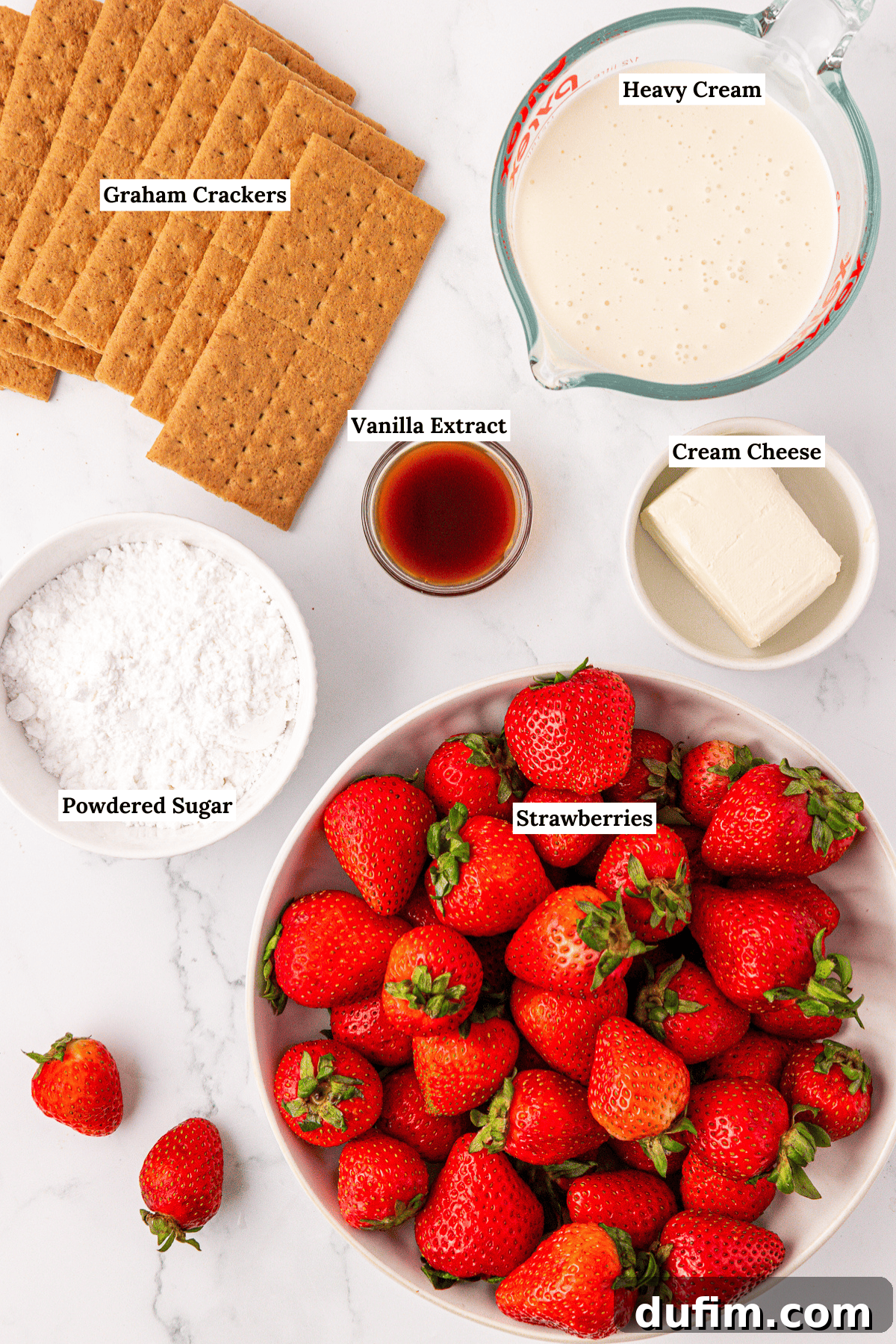 No-Bake Strawberry Dream 5 Top-down view of ingredients on a white surface: graham crackers, a bowl of strawberries, a block of cream cheese, a measuring cup of heavy cream, vanilla extract in a small bowl, and a bowl of powdered sugar.