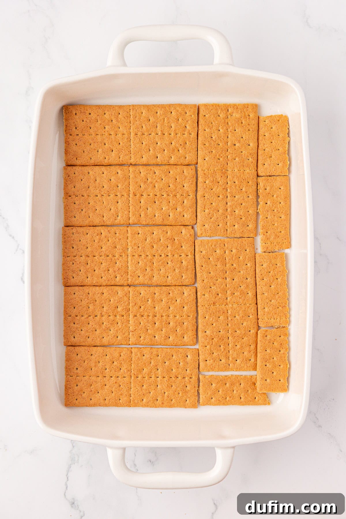 No-Bake Strawberry Dream 6 A white rectangular baking dish lined with pieces of graham crackers arranged to cover the bottom, set on a white marble surface.