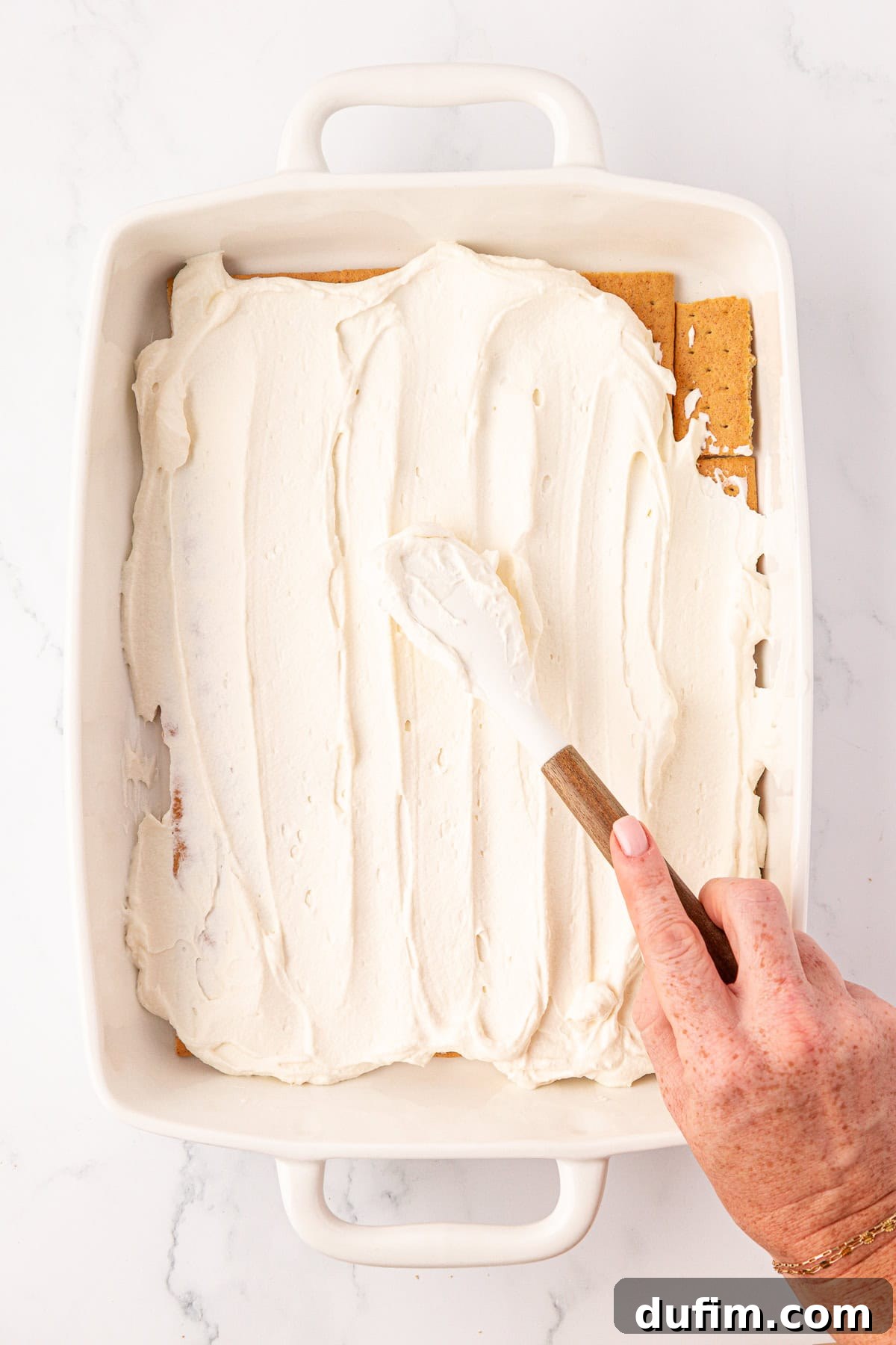 No-Bake Strawberry Dream 7 A hand uses a spatula to spread creamy white frosting over layered graham crackers in a rectangular white baking dish on a marble surface.