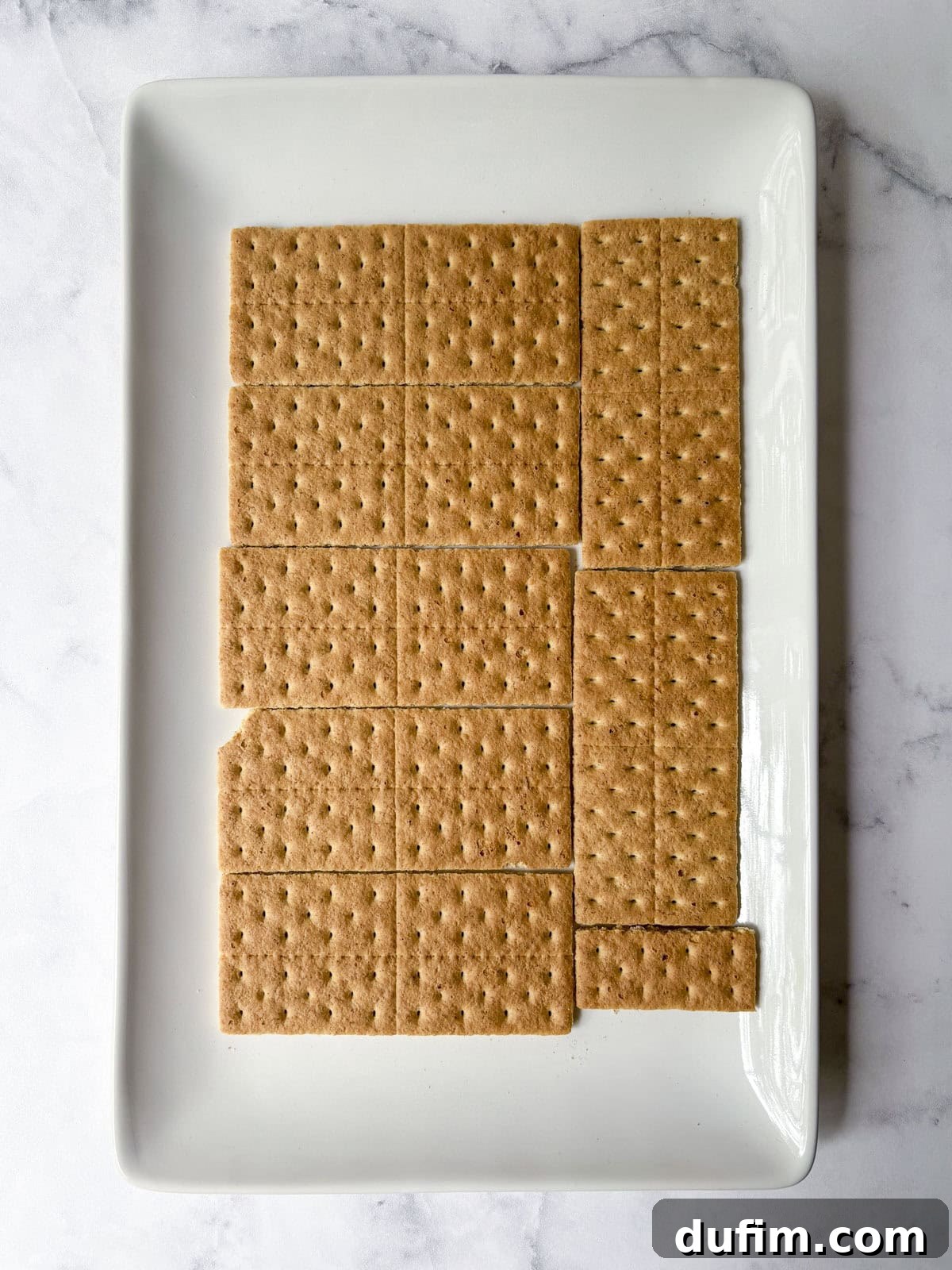 No-Bake Strawberry Dream 8 A white rectangular plate holds 14 rectangular graham crackers neatly arranged, with one cracker missing a corner and one small, separate square piece on the bottom right. The background is a light marble surface.