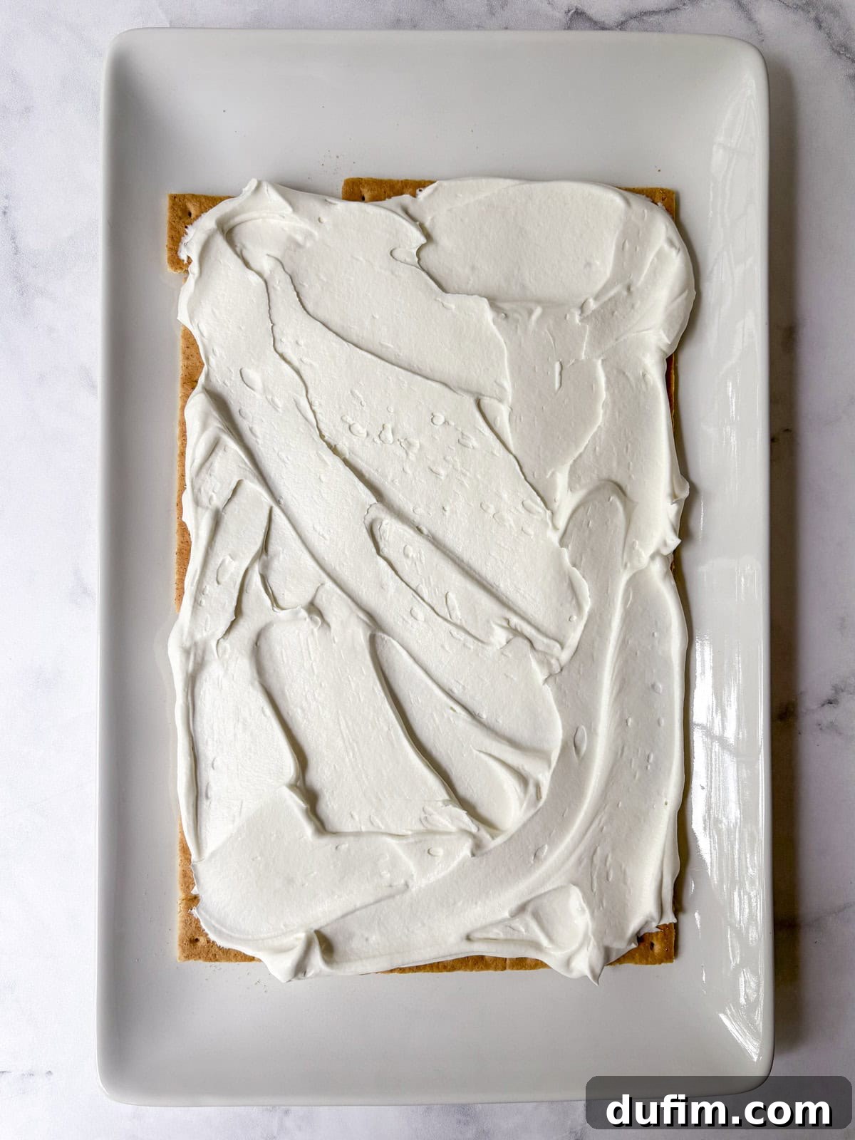 No-Bake Strawberry Dream 9 A rectangular graham cracker topped with a thick, even layer of white frosting sits on a white rectangular plate against a marble background.