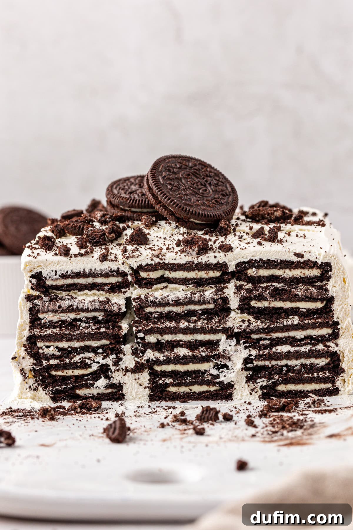 A layered cookies and cream icebox cake with visible rows of chocolate sandwich cookies and cream, topped with more cookies and crumbled pieces, on a white surface.