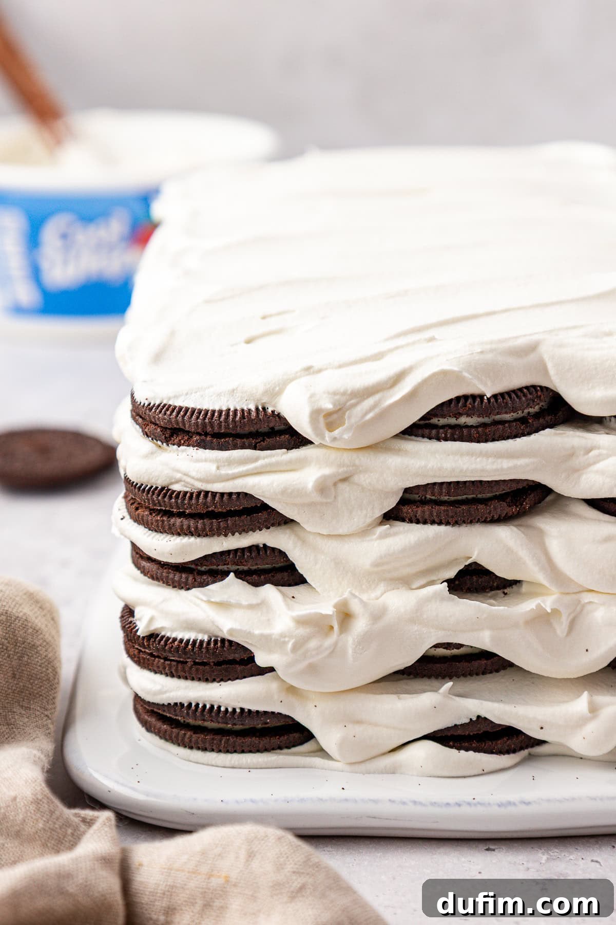 A close-up of a layered dessert made with chocolate sandwich cookies and whipped cream, forming a rectangular cake shape on a white surface. A container of whipped topping and a cookie are in the background.