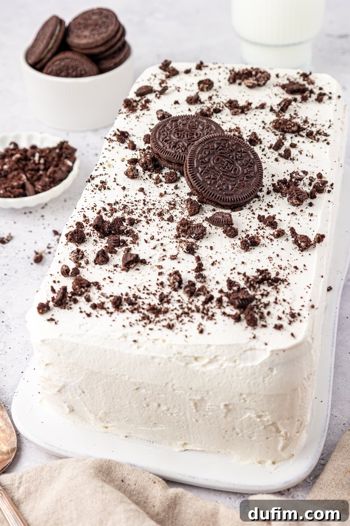 Rectangular cookies and cream ice cream cake topped with whole and crushed chocolate sandwich cookies, placed on a white platter. Bowls of cookies and cookie crumbs are in the background.