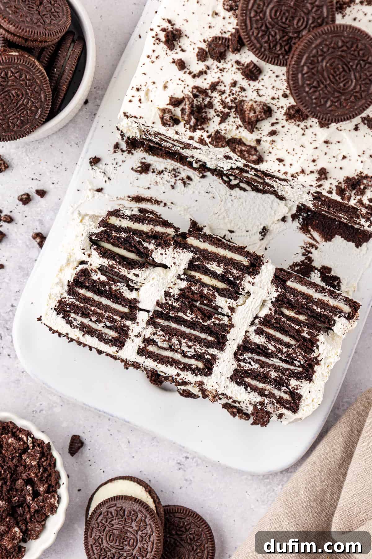 A sliced Oreo icebox cake on a white platter shows layers of chocolate cookies and whipped cream, with whole and crushed Oreo cookies scattered nearby on a light countertop.