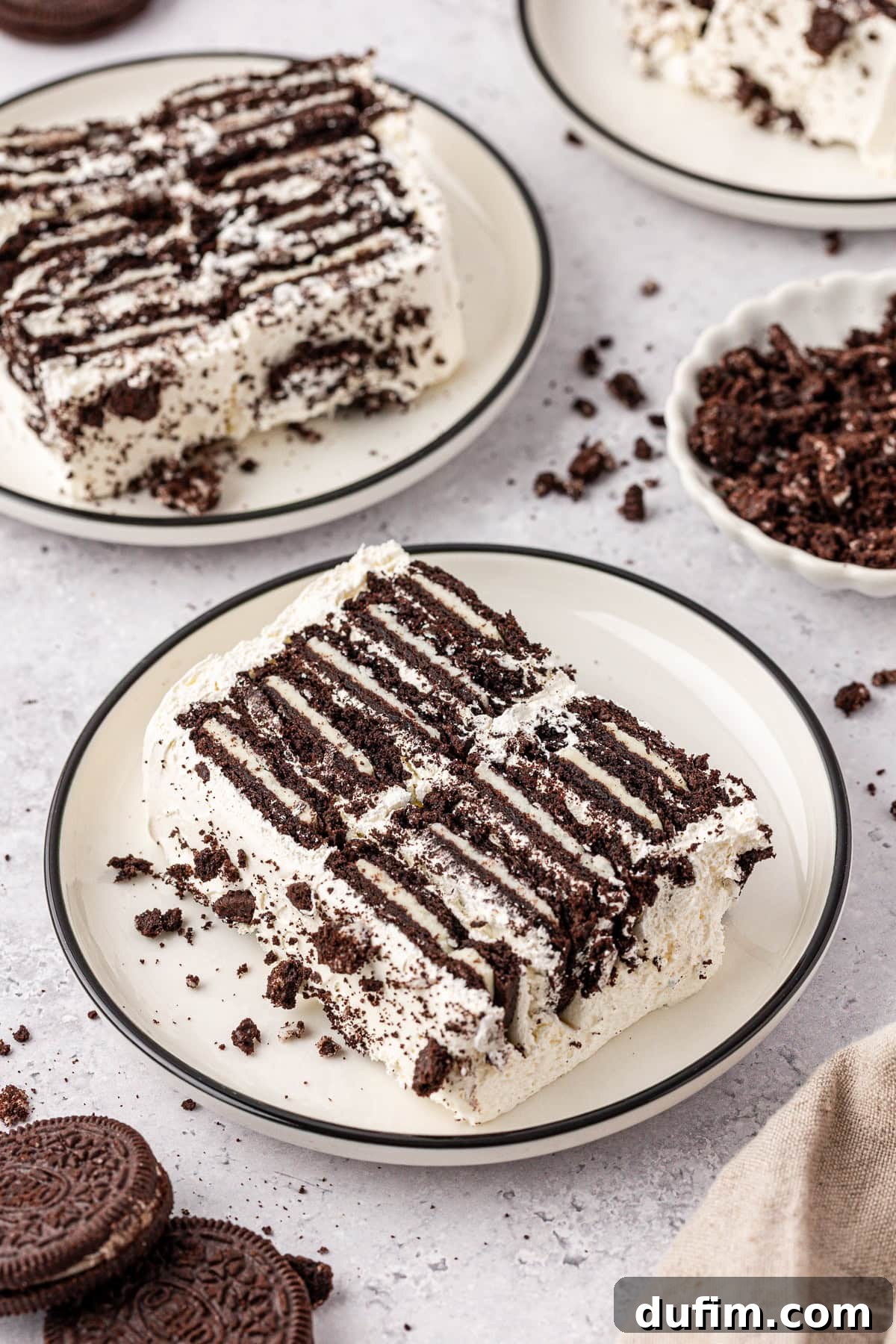Two slices of cookies and cream icebox cake are served on a plate, featuring layers of chocolate cookies and whipped cream. Crumbled cookies are scattered around, with extra plates and whole cookies in the background.