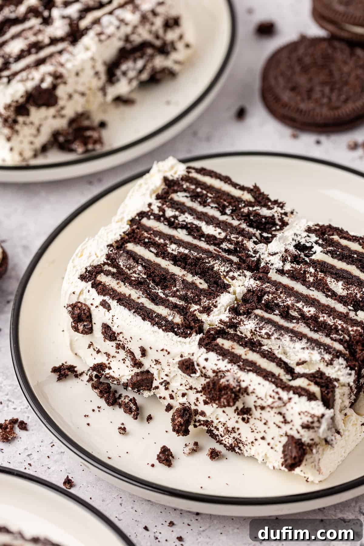 A close-up of a dessert made with layers of chocolate cookies and cream, sliced and served on a plate. Crumbled cookie pieces are scattered on top and around the dessert.