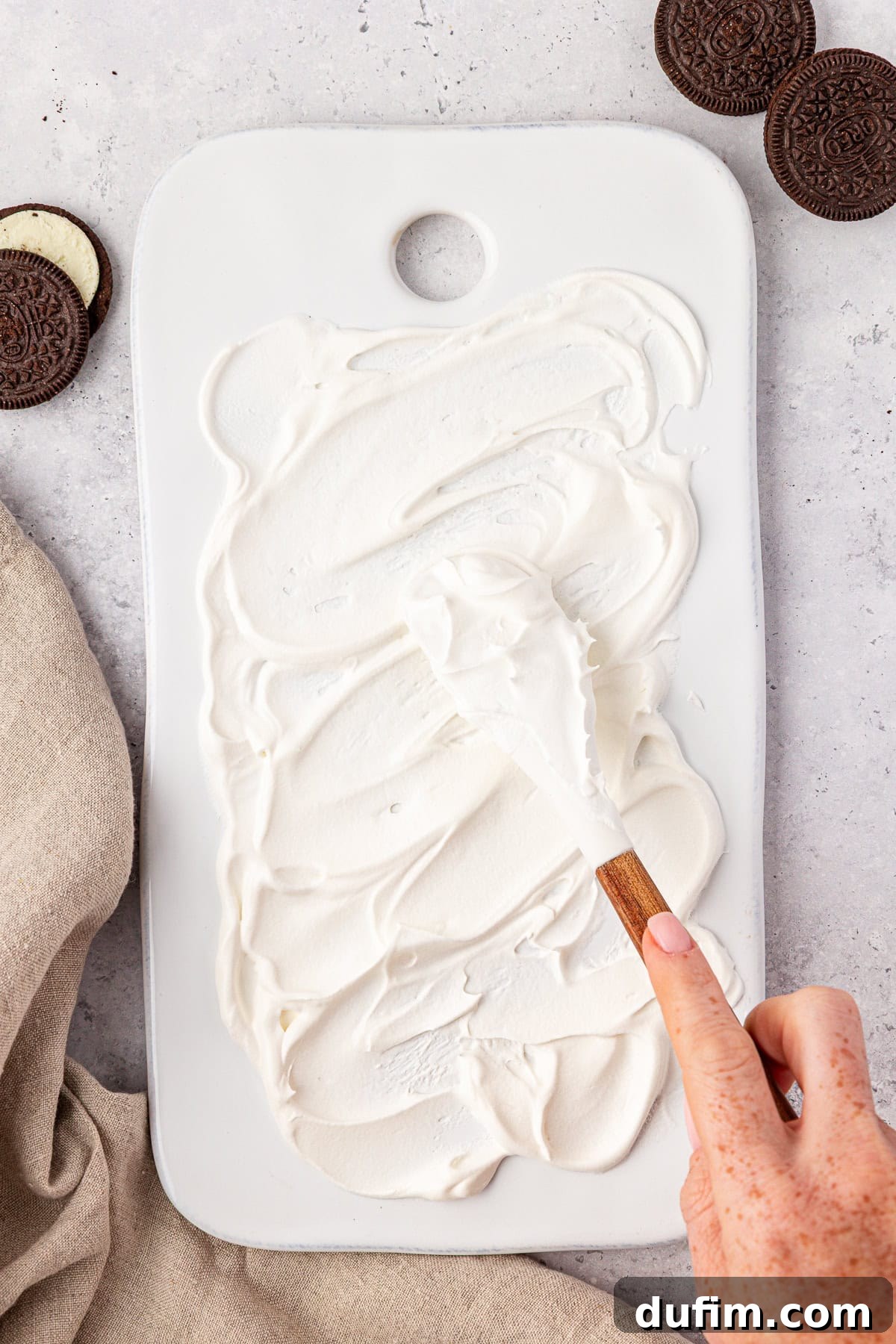 A hand spreads creamy white frosting on a rectangular white cutting board with a wooden-handled spatula. Chocolate sandwich cookies and a beige cloth are nearby on a light surface.