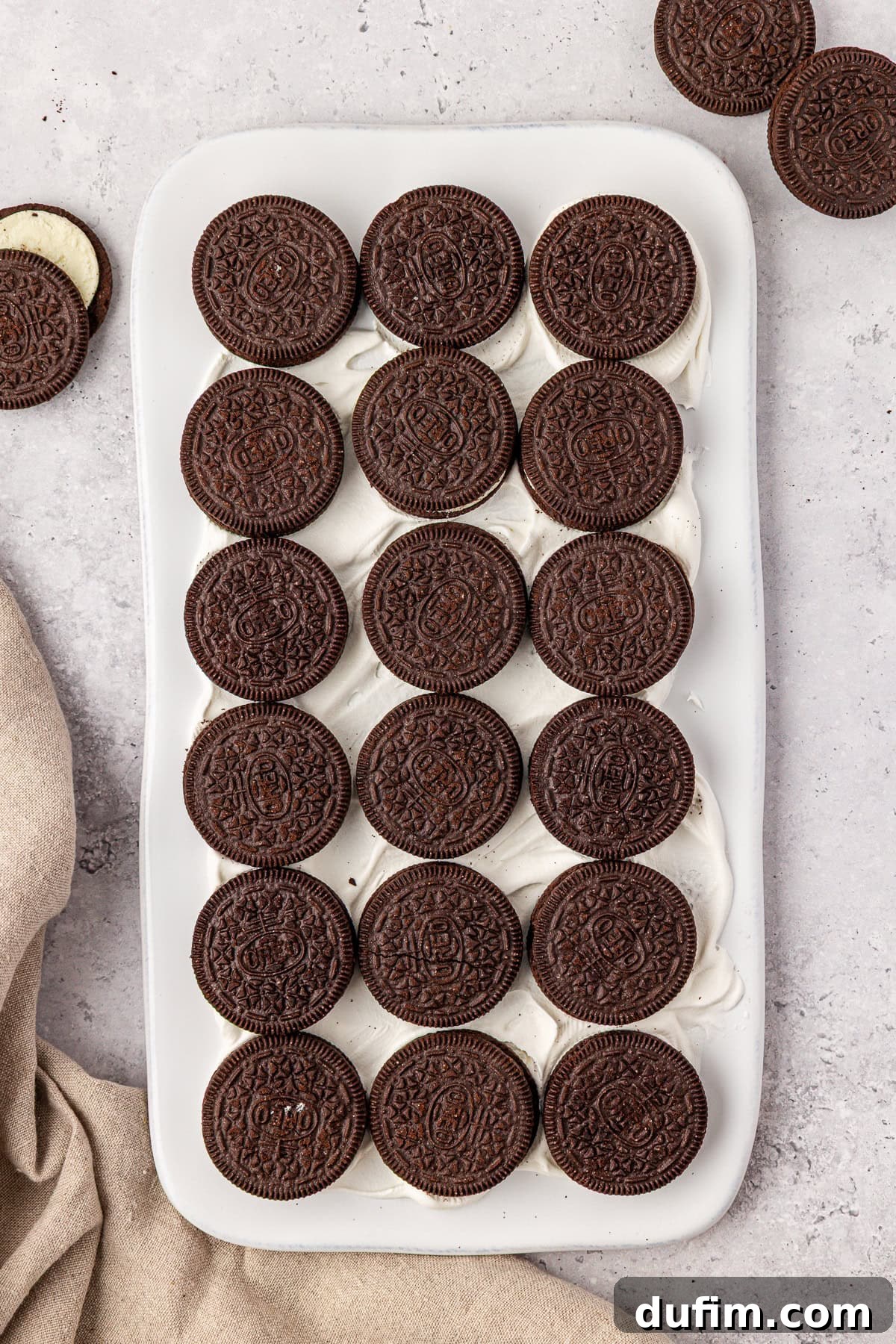 A white rectangular dish filled with rows of chocolate sandwich cookies arranged neatly on top of a layer of creamy white filling, set on a light gray countertop with a beige cloth nearby.