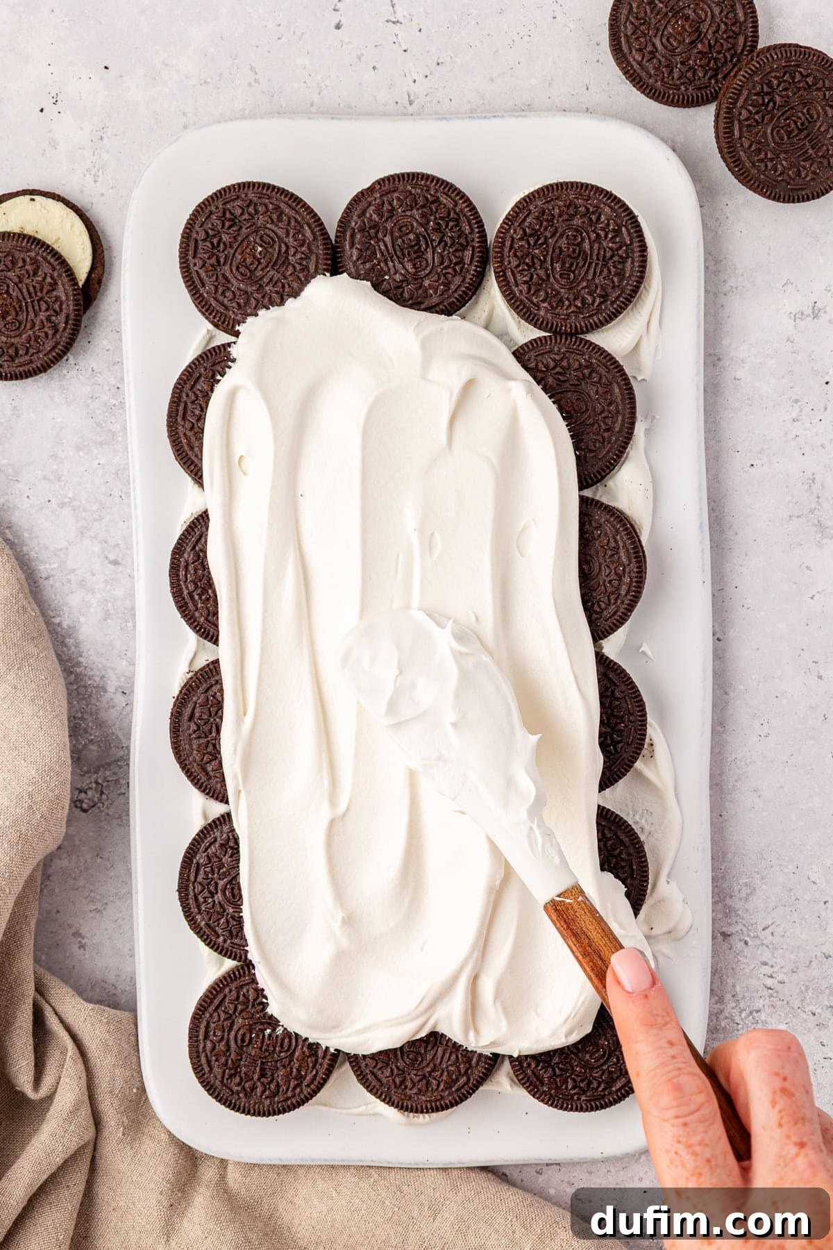 A hand spreads whipped cream over a layer of chocolate sandwich cookies arranged in a white rectangular dish, with extra cookies and a beige cloth nearby on a light surface.