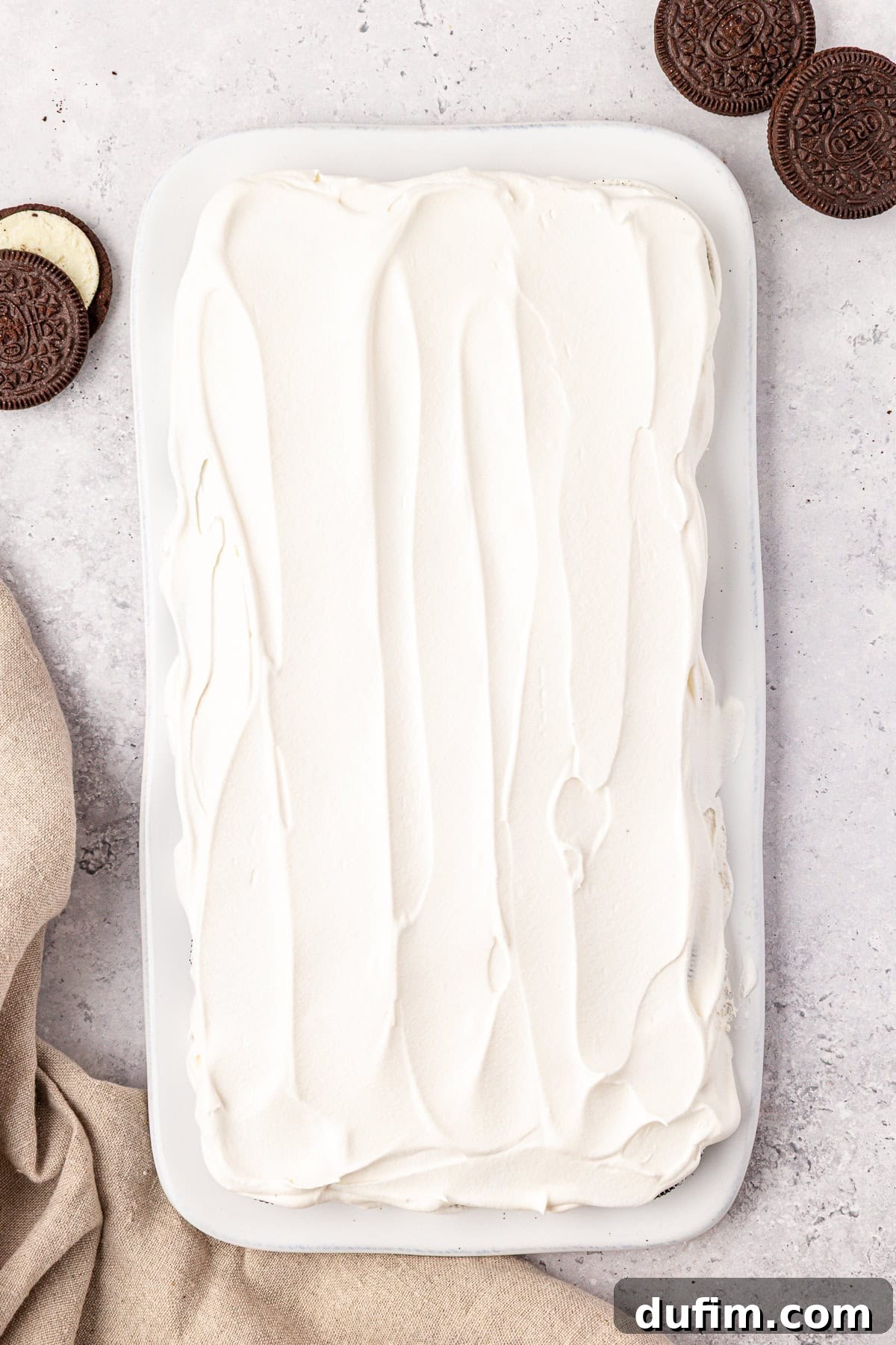 A rectangular dish filled with a dessert covered in smooth, white whipped topping. Chocolate sandwich cookies are placed nearby on a light textured surface, with a beige cloth partially visible in the corner.