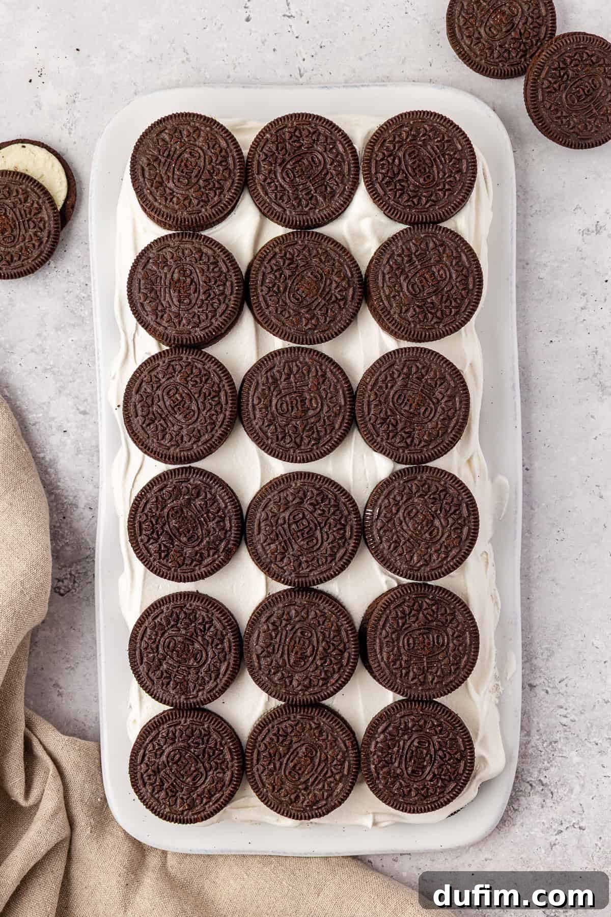 A rectangular tray of dessert topped with neatly arranged whole chocolate sandwich cookies, placed on a light surface with a beige cloth and a few extra cookies nearby.