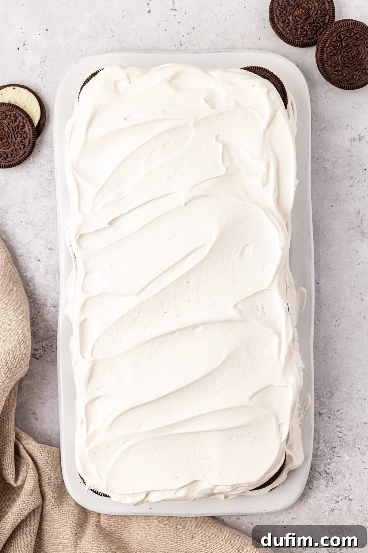 A rectangular dessert topped with smooth white whipped cream sits on a white tray, with chocolate sandwich cookies and a beige cloth beside it on a light marble surface.