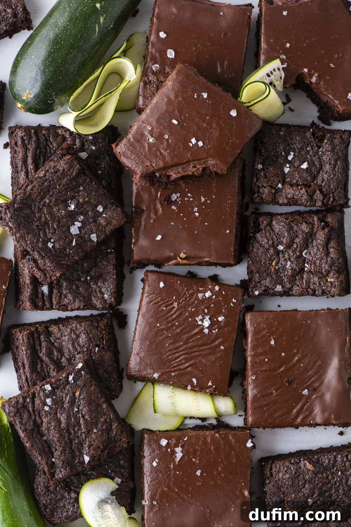Hidden Harvest Brownies 2 A top view of chocolate brownies, some with glossy frosting and a sprinkle of sea salt, arranged with fresh zucchini and zucchini ribbons on a white surface.