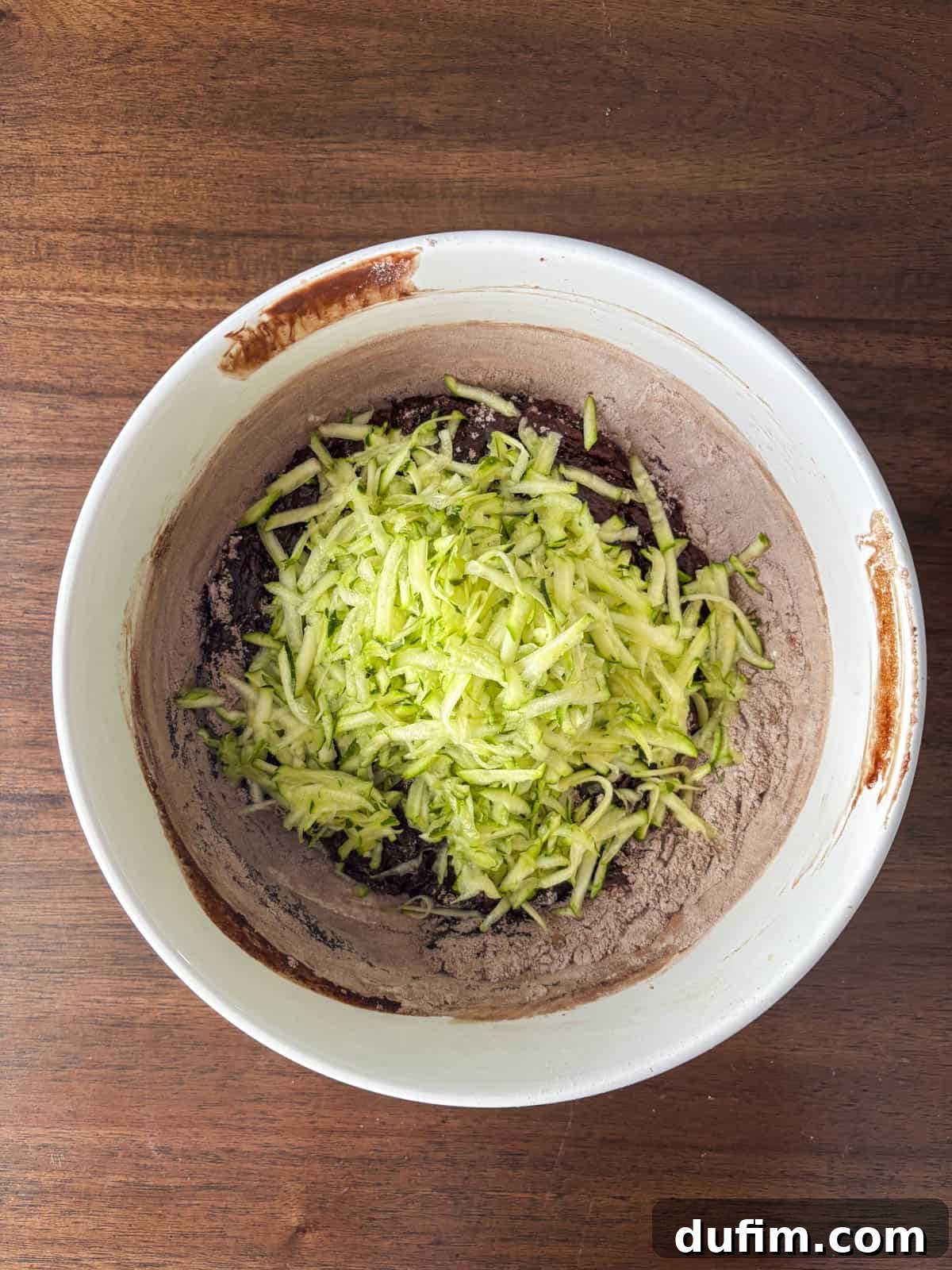 Hidden Harvest Brownies 11 A white mixing bowl with chocolate cake batter and a pile of freshly grated zucchini on top, sitting on a wooden surface.
