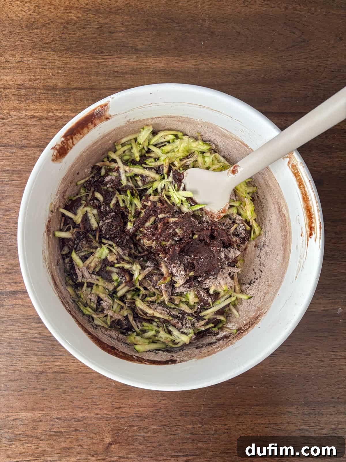 Hidden Harvest Brownies 12 A white mixing bowl filled with brownie batter and shredded zucchini sits on a wooden surface. A white spatula is resting in the bowl, ready to stir the ingredients together.