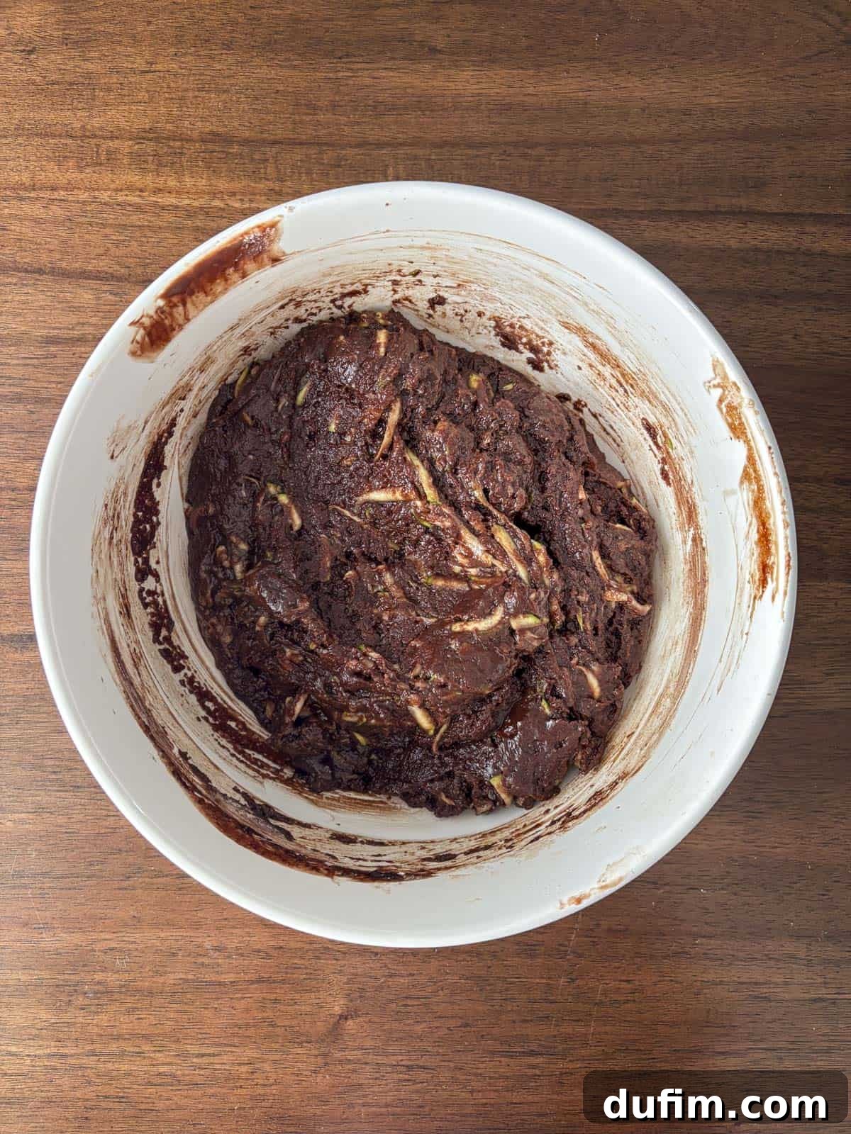 Hidden Harvest Brownies 13 A white mixing bowl on a wooden surface contains thick, dark chocolate batter with visible shreds of zucchini mixed in.