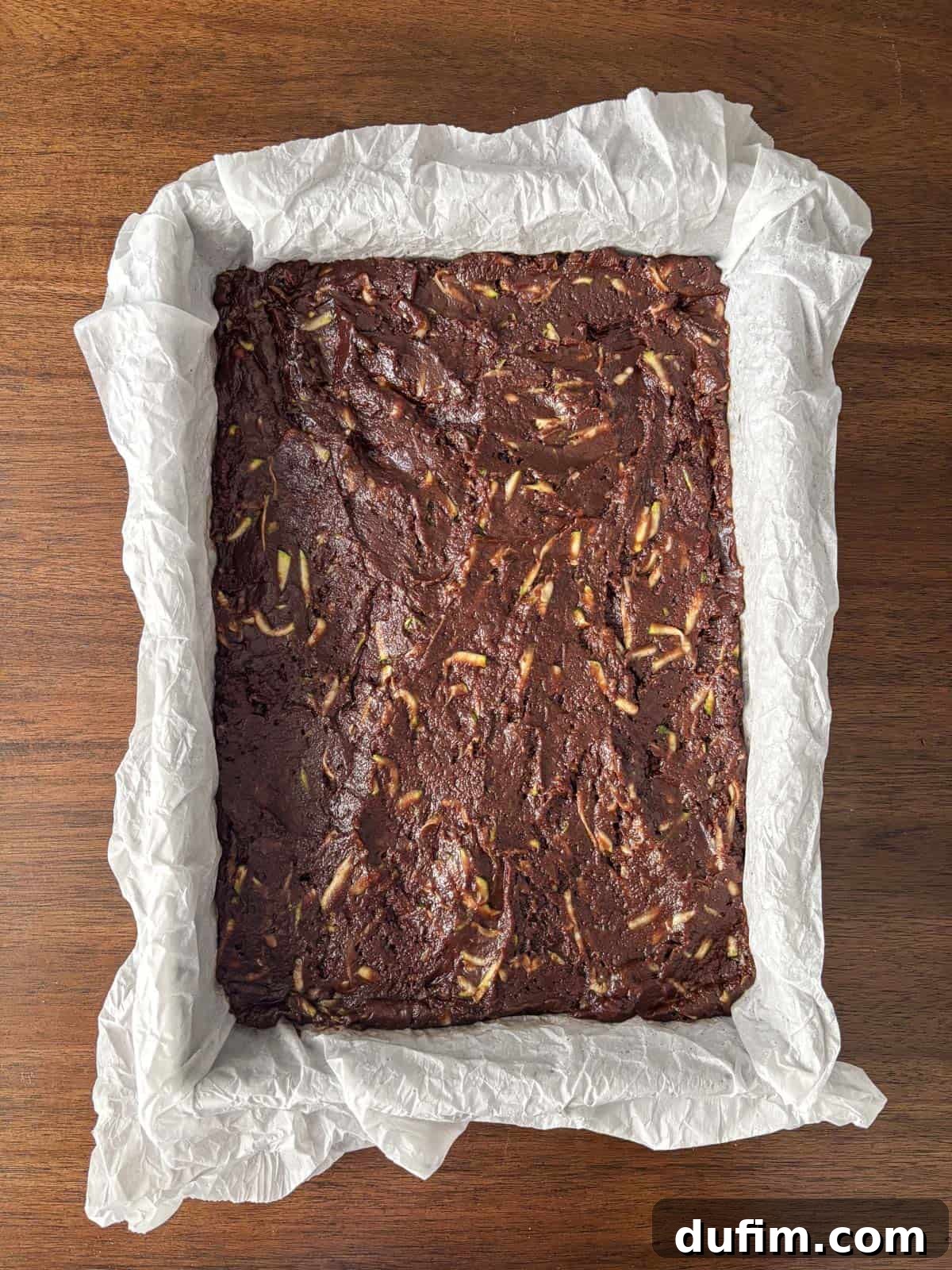 Hidden Harvest Brownies 14 A rectangular baking pan lined with crinkled parchment paper holds unbaked chocolate brownie batter with visible almond slivers, placed on a wooden surface.