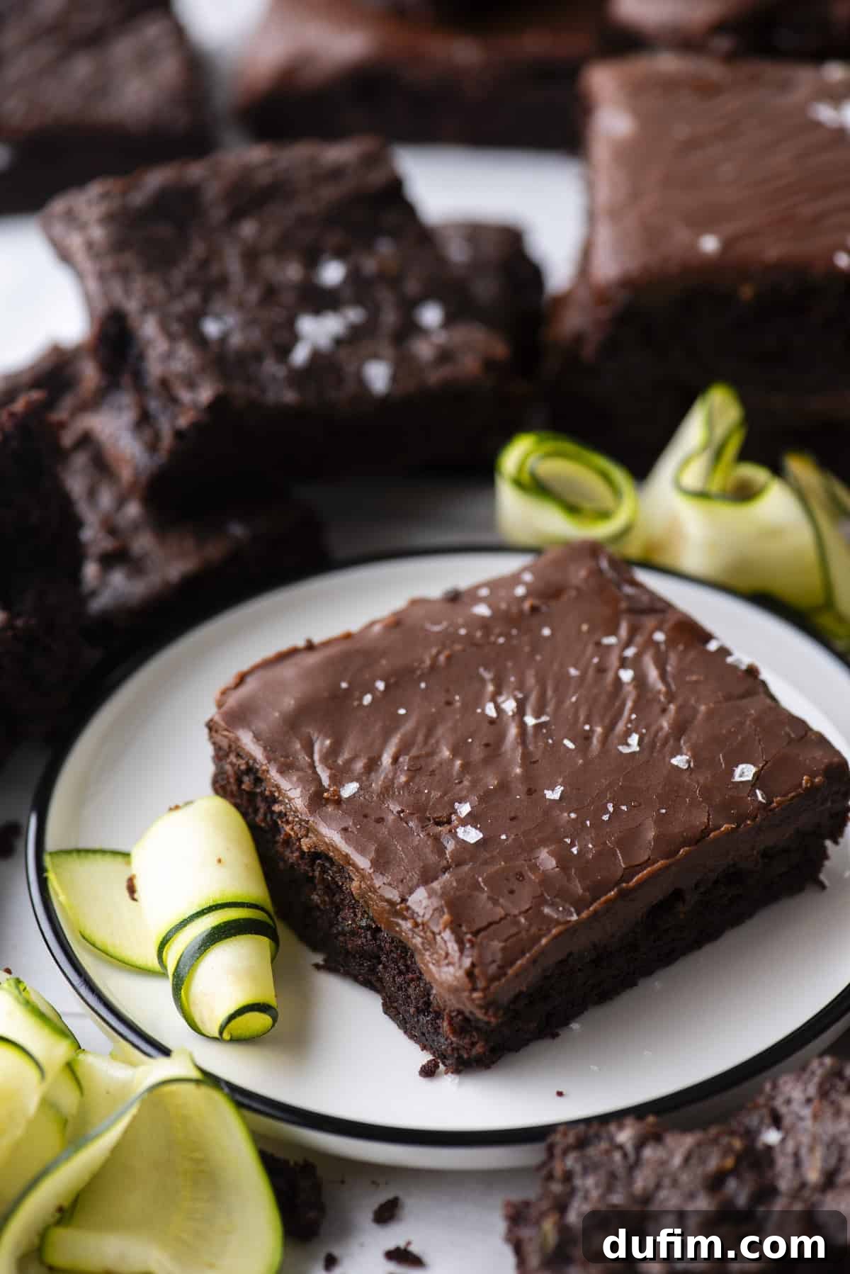 Hidden Harvest Brownies 16 A chocolate brownie topped with flaky sea salt sits on a white plate, surrounded by chocolate brownies and garnished with fresh zucchini ribbons.
