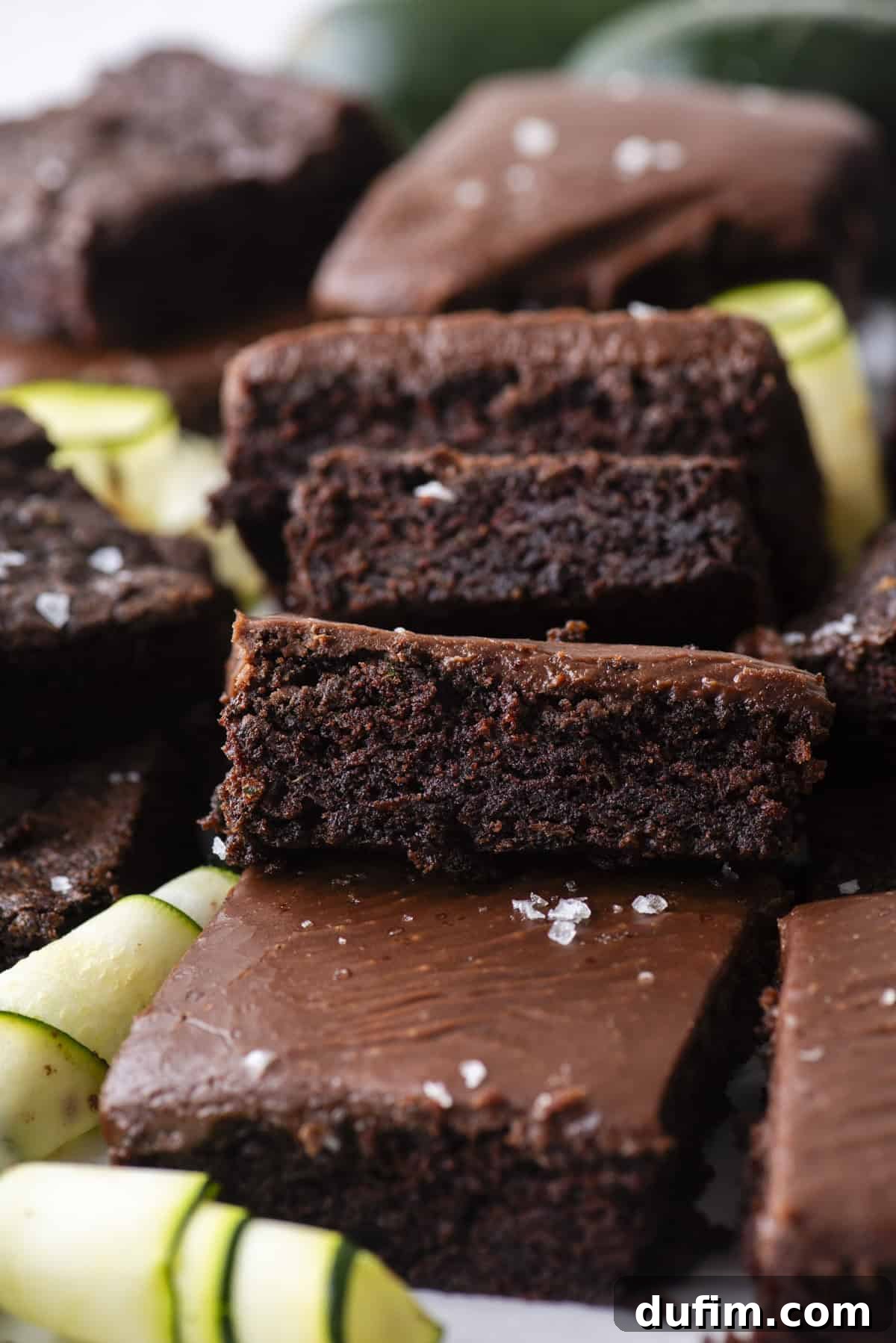 Hidden Harvest Brownies 17 A close-up of rich, fudgy chocolate brownies stacked on top of each other, sprinkled with flaky sea salt. Slices of fresh zucchini are scattered around the brownies.