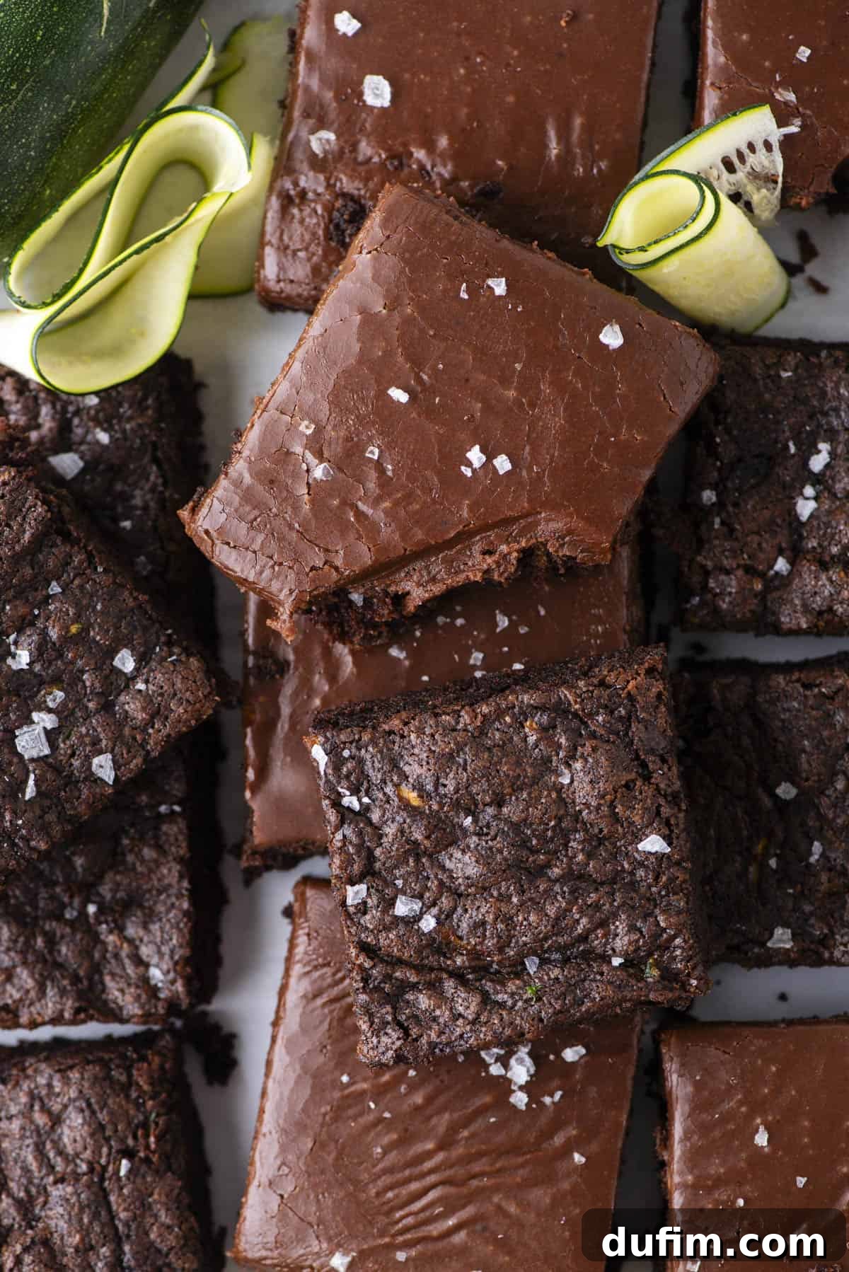 Hidden Harvest Brownies 18 A close-up of chocolate brownies topped with flaky sea salt, some with chocolate frosting. Zucchini ribbons are placed in the corner, hinting at a possible ingredient in the brownies.