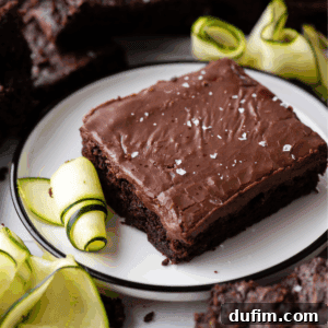 A chocolate zucchini brownie topped with flaky sea salt is served on a white plate, garnished with thinly sliced zucchini ribbons. Additional brownies and zucchini slices are visible in the background.