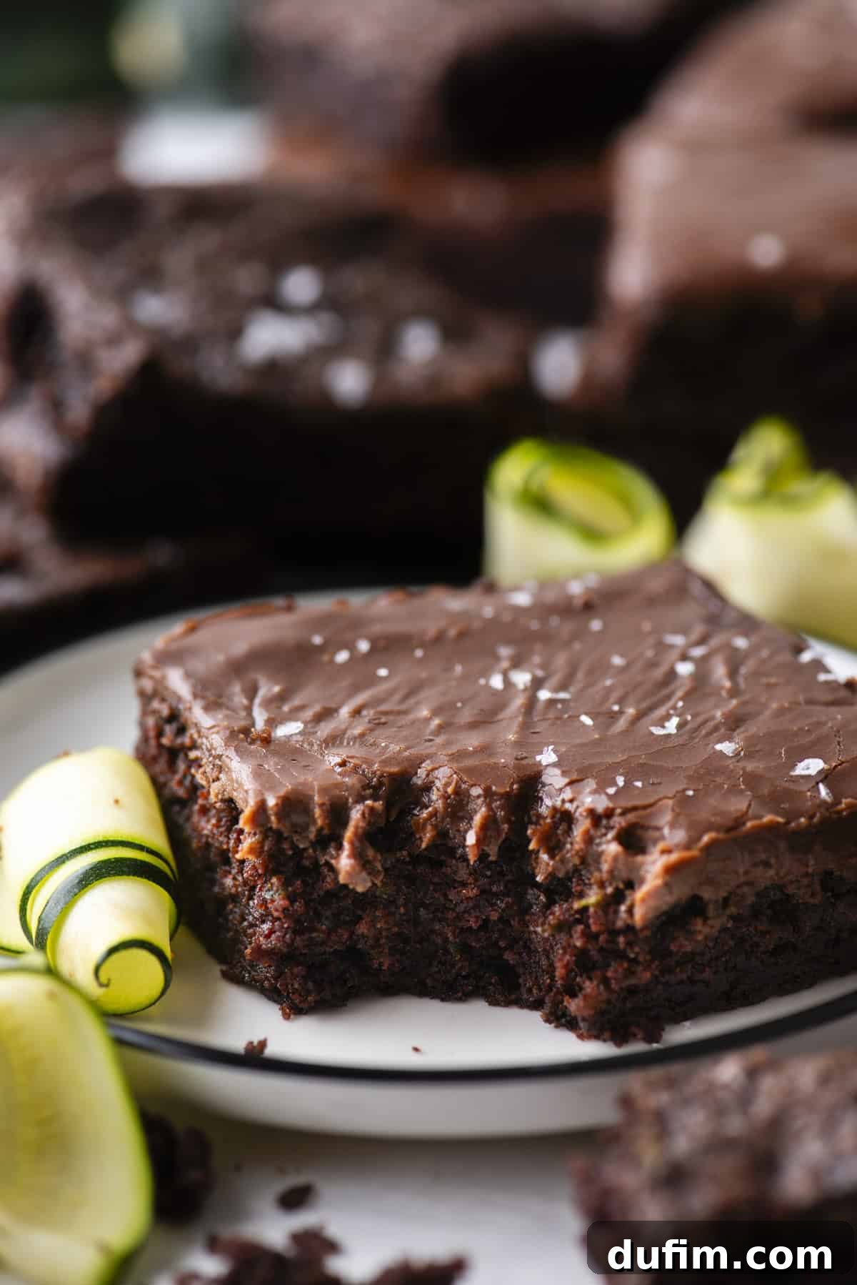 Hidden Harvest Brownies 3 A close-up of a chocolate frosted brownie with a bite taken out, garnished with sea salt flakes and zucchini ribbons, served on a white plate with more brownies in the background.