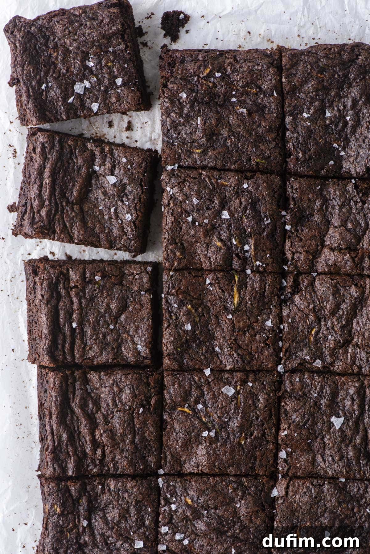 Hidden Harvest Brownies 4 A batch of chocolate brownies cut into squares on parchment paper. Two brownies are stacked in the top left corner. The brownies are topped with a sprinkle of flaky sea salt.