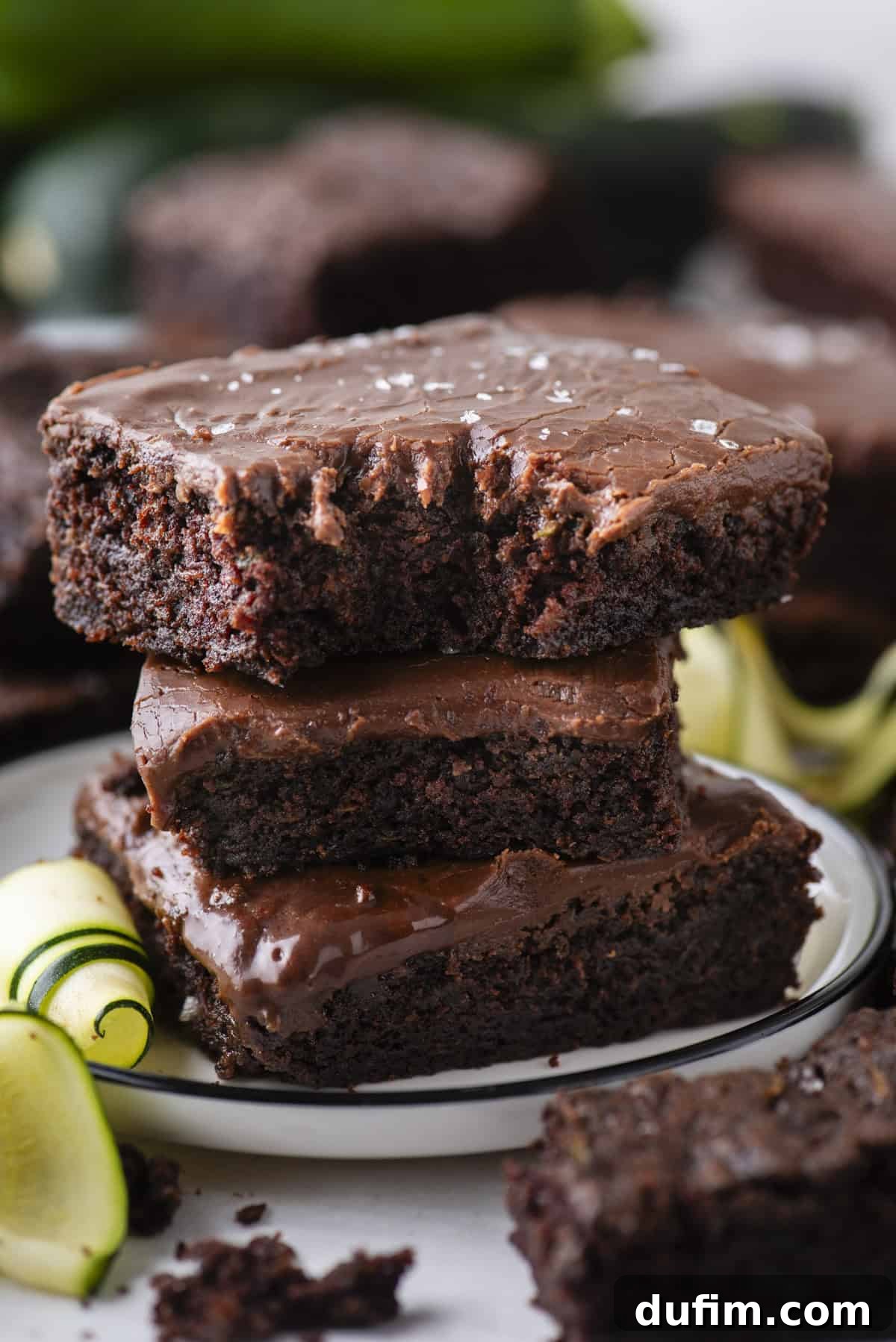 Hidden Harvest Brownies 6 A stack of three chocolate brownies with glossy frosting, sprinkled with sea salt, sits on a white plate. Slices of zucchini are beside the brownies. The background shows more brownies.