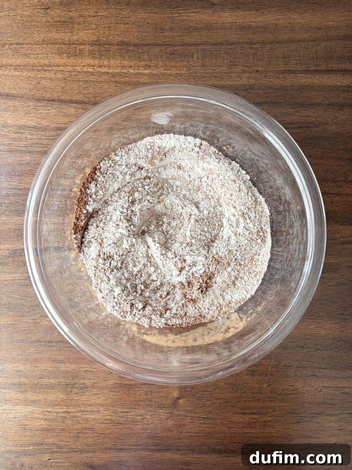 Hidden Harvest Brownies 7 A glass bowl filled with a mixture of dry ingredients, including flour and cocoa powder, sits on a wooden surface.