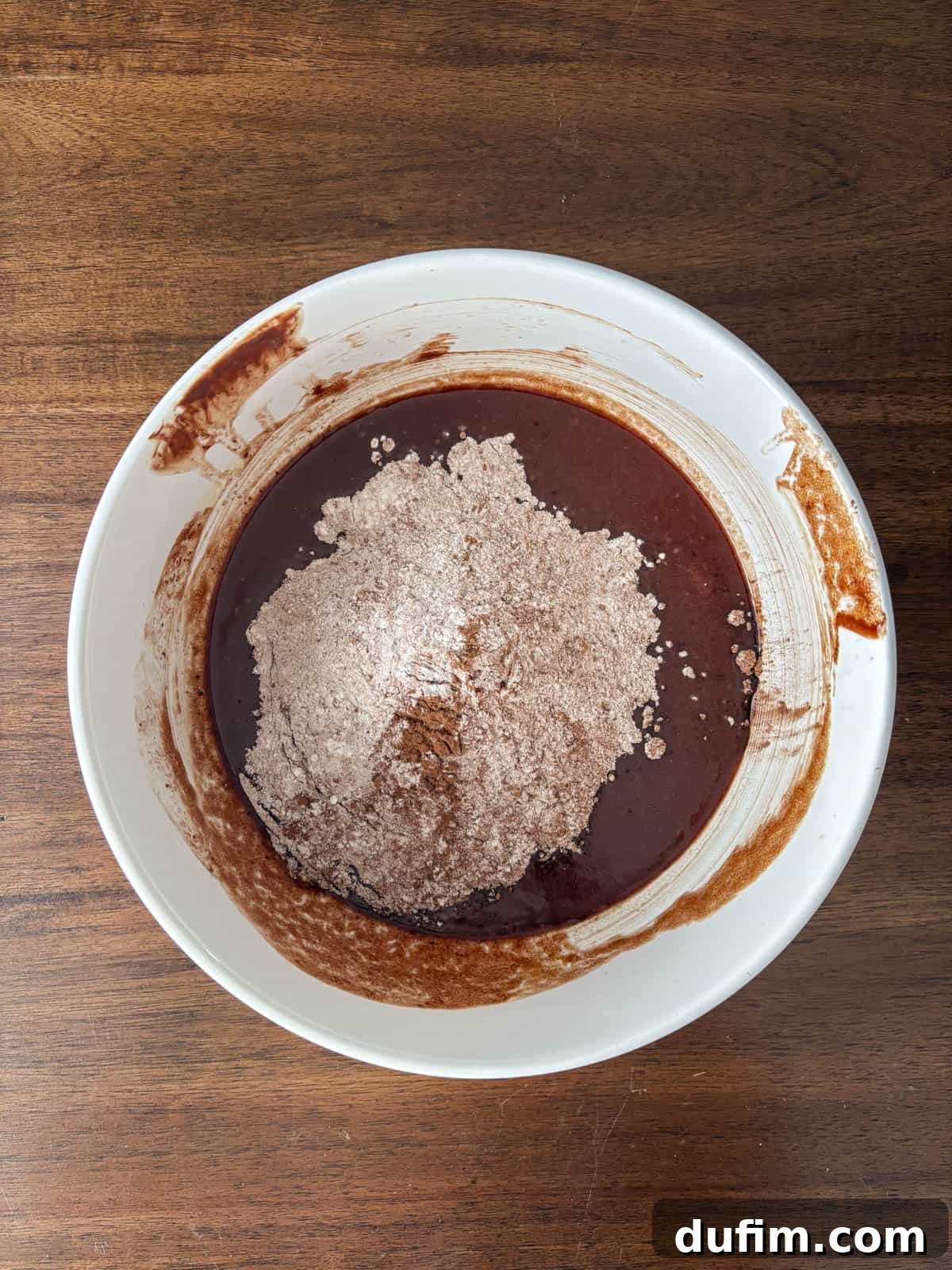Hidden Harvest Brownies 10 A white bowl on a wooden surface contains brownie batter with a mound of dry cocoa powder and flour in the center, partially mixed into the wet ingredients.