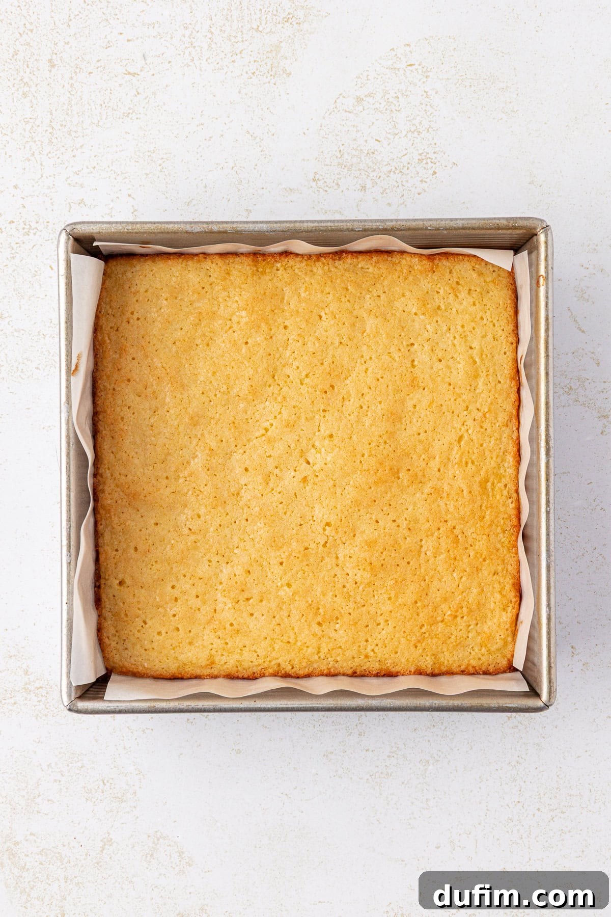 Zesty Lemon Brownies 11 A golden-brown, freshly baked square cake sits in a parchment-lined metal baking pan on a light, textured surface.