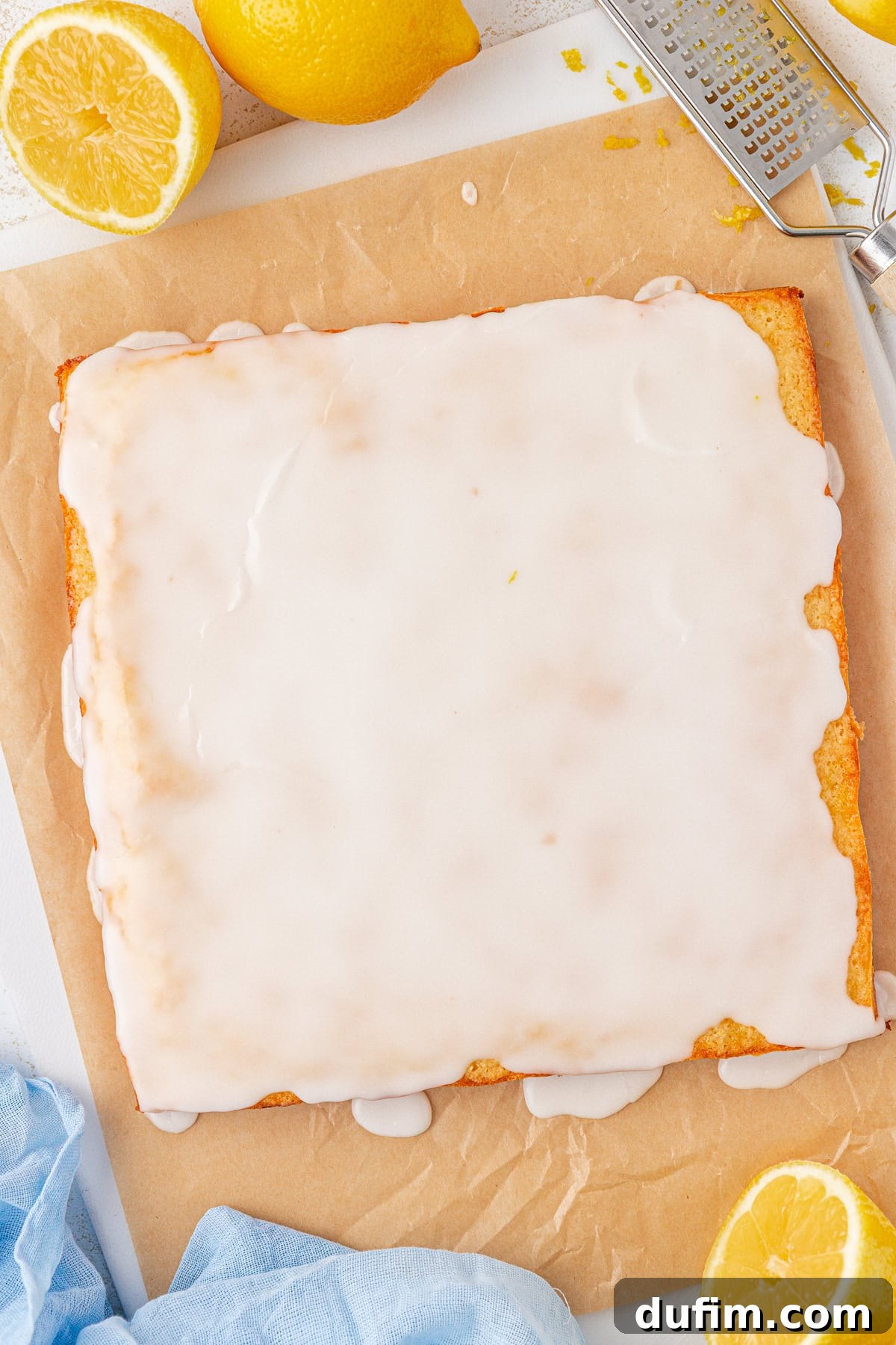 Zesty Lemon Brownies 13 A rectangular lemon cake with white glaze sits on parchment paper, surrounded by fresh lemons and a grater. Some glaze drips down the sides of the cake. A blue cloth is partially visible in the corner.
